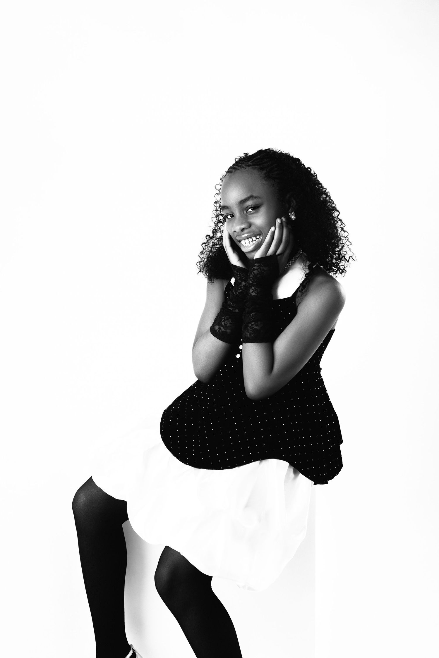 Black and white portrait of a smiling young woman in a black top and white skirt, posing with hands on cheek.