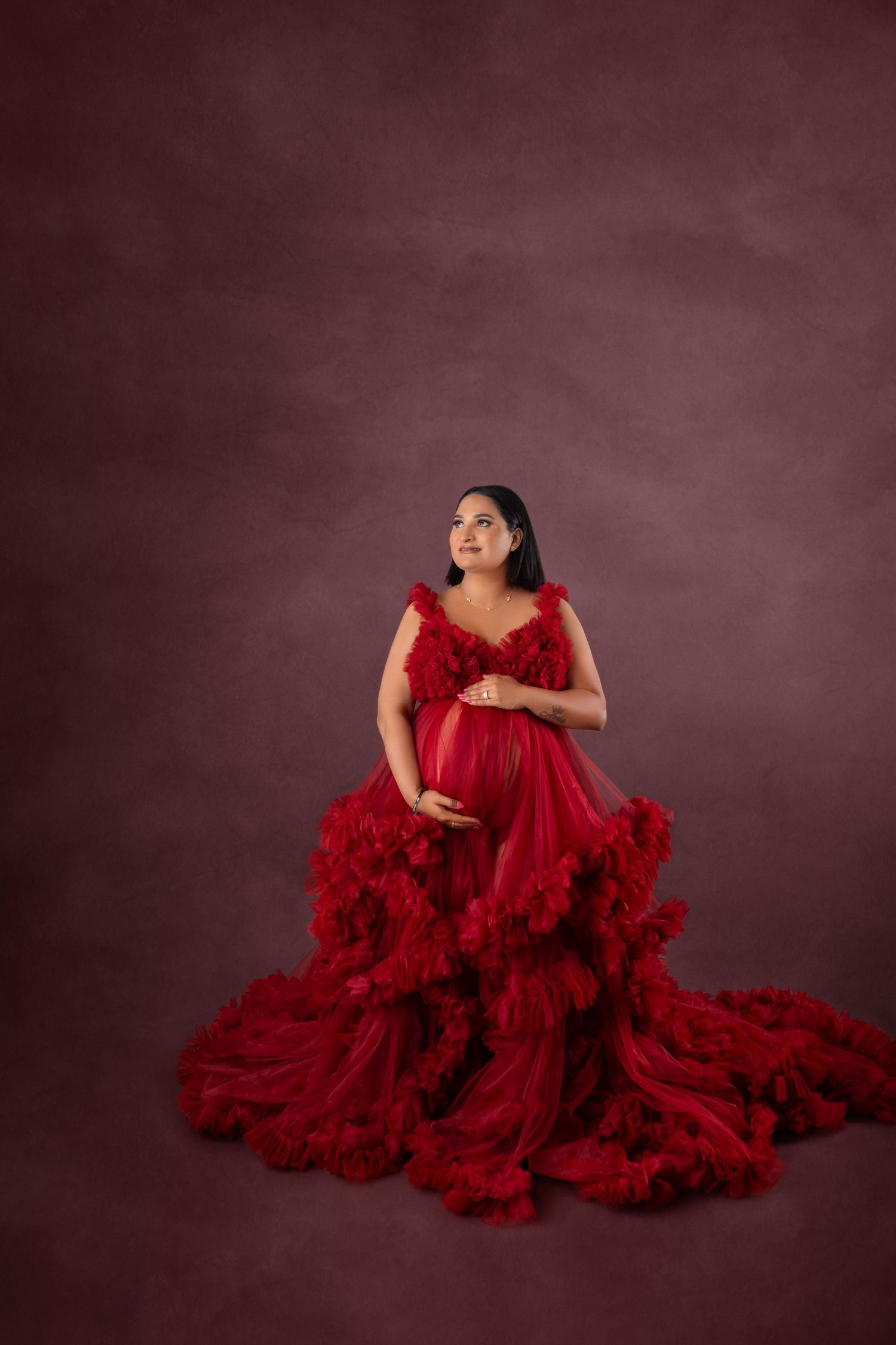 Pregnant woman in elegant red ruffled tulle gown sitting gracefully against a moody dark red studio background.