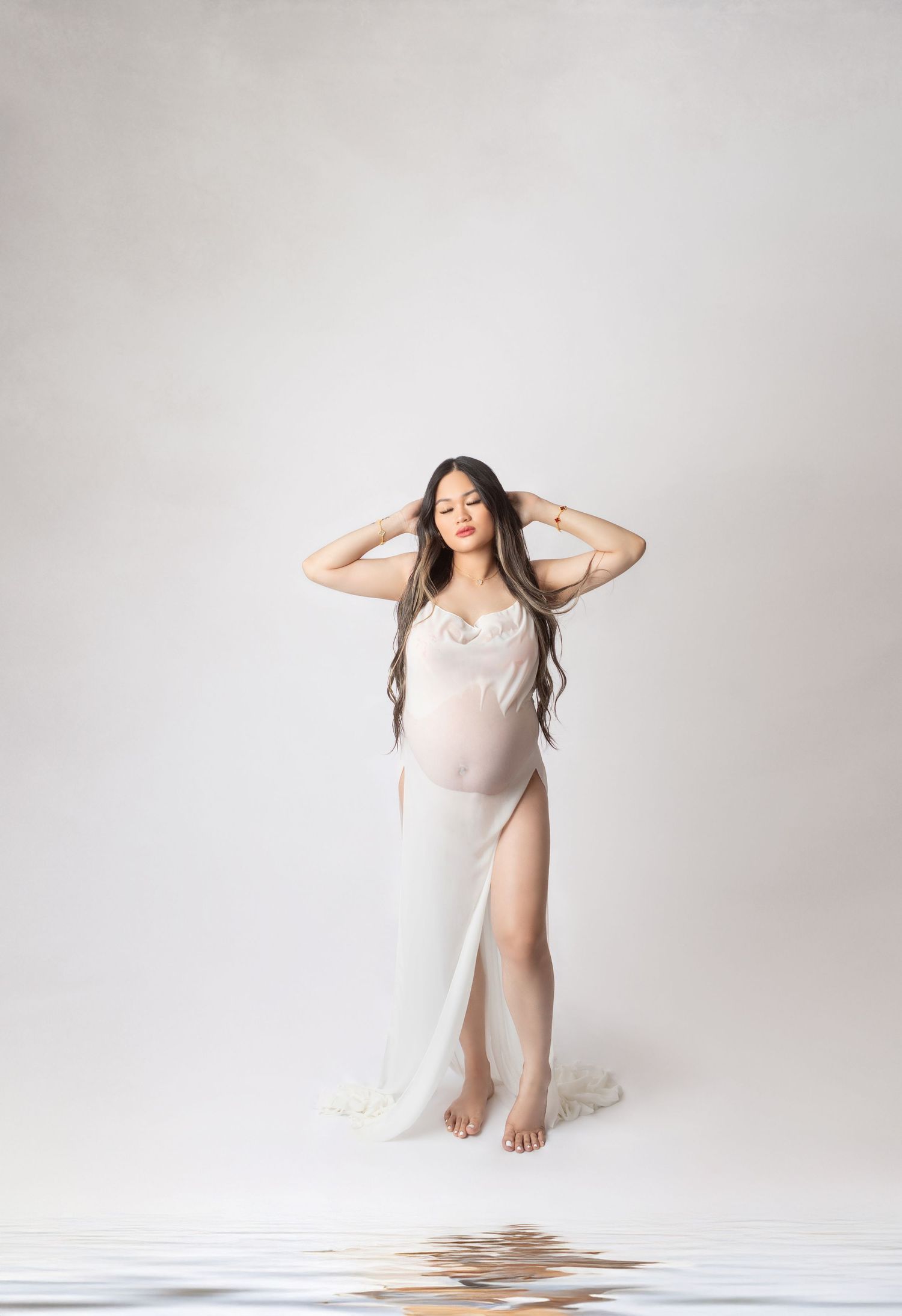 Pregnant woman in sheer white gown posing gracefully in studio with water reflection.
