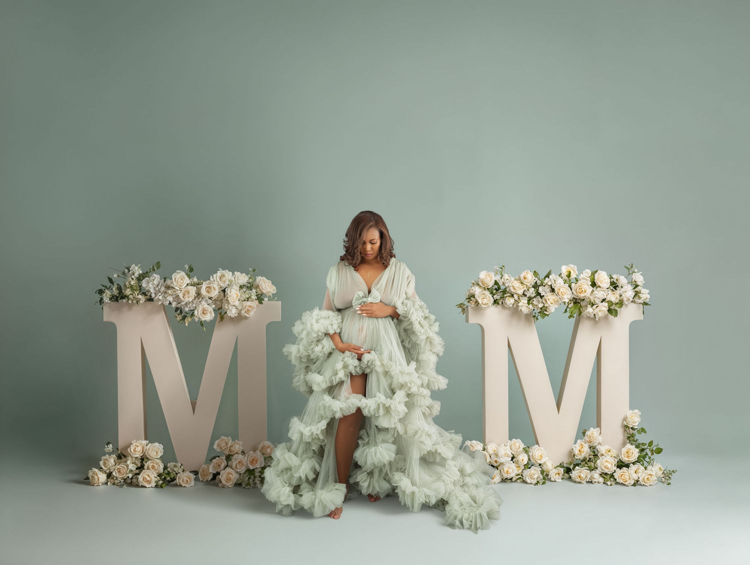 Pregnant woman in mint ruffled gown poses between large floral M letters for maternity photo shoot.