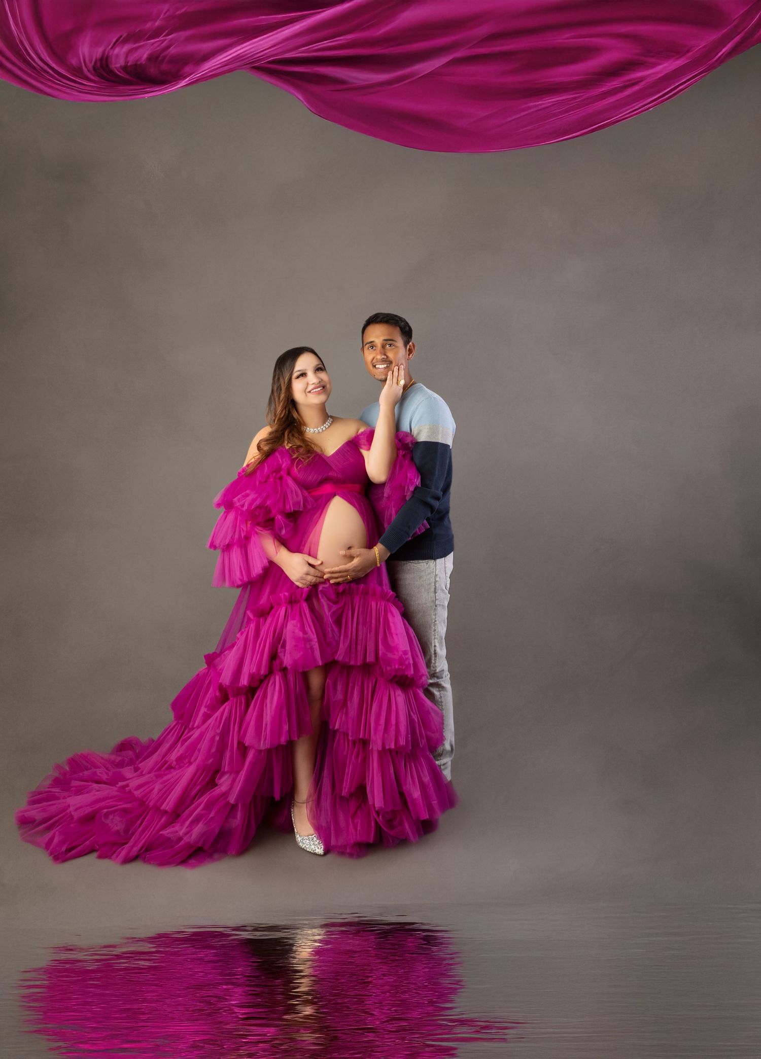 Pregnant woman in magenta ruffle gown poses with partner in studio maternity shoot with flowing fabric.