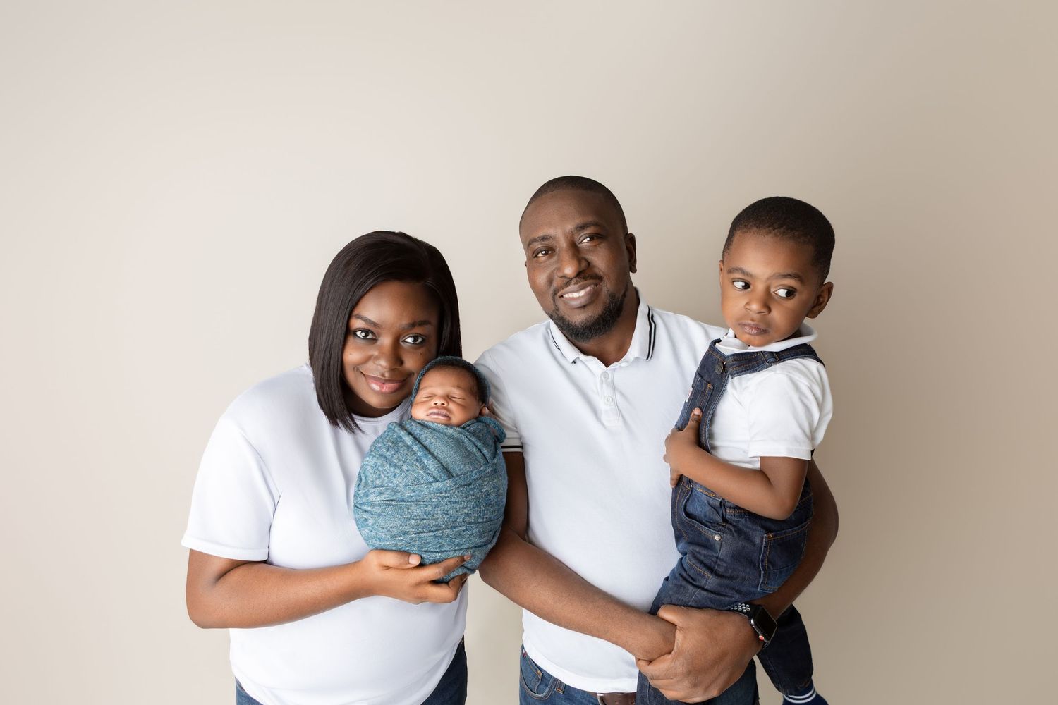 A happy family of four poses for a portrait, with parents holding their newborn and toddler against a neutral background.