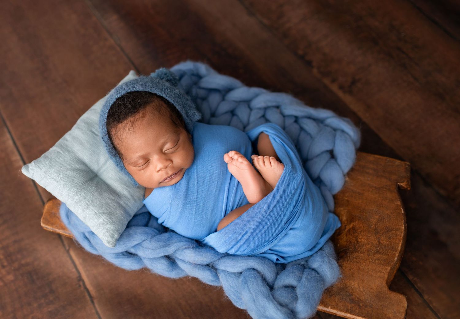 Newborn baby wrapped in blue swaddle sleeping peacefully on chunky knit blanket in wooden bowl prop.
