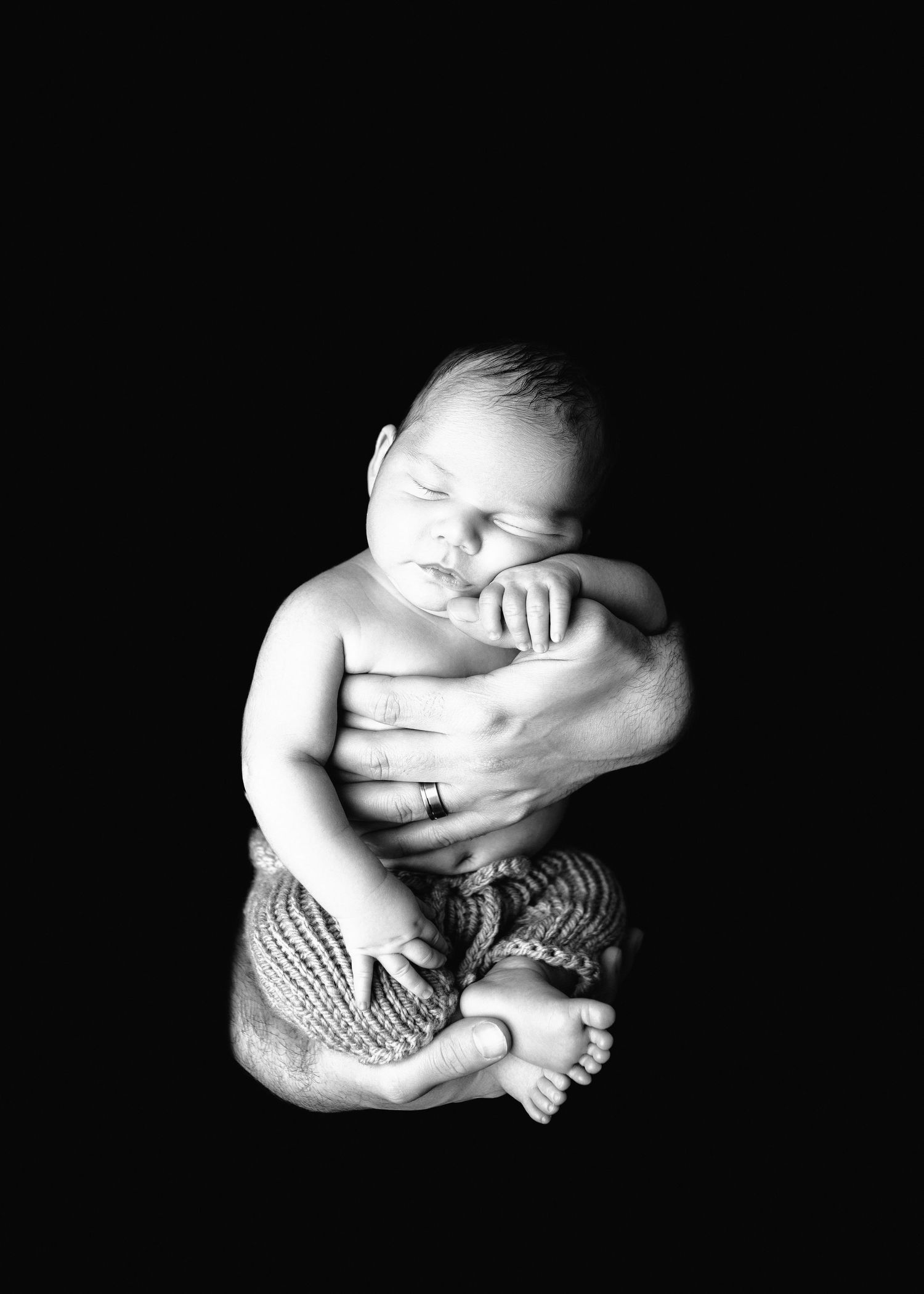 Black and white newborn photo of sleeping baby cradled in adult hands against dark background.