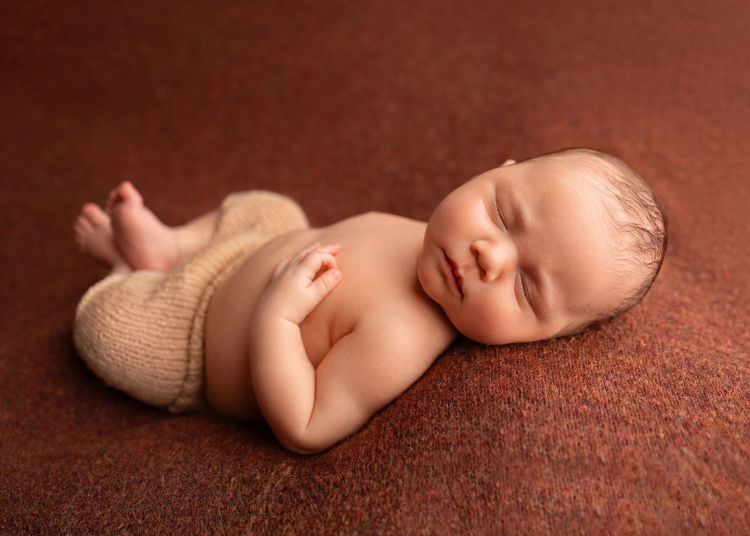 Newborn baby in beige knit shorts sleeping peacefully on a warm brown textured blanket.