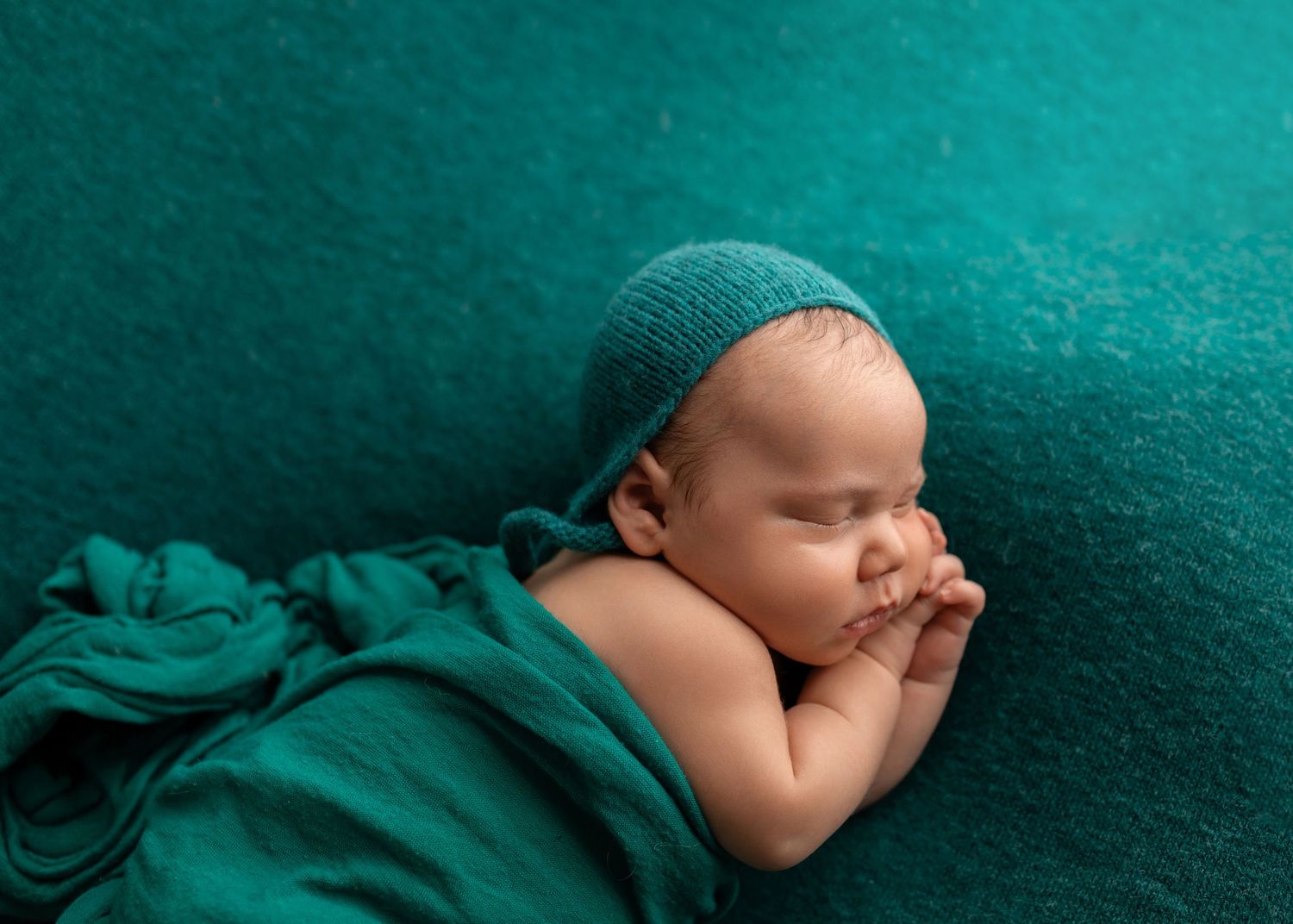 Peaceful newborn baby in teal knit bonnet wrapped in teal fabric, sleeping with hands together.