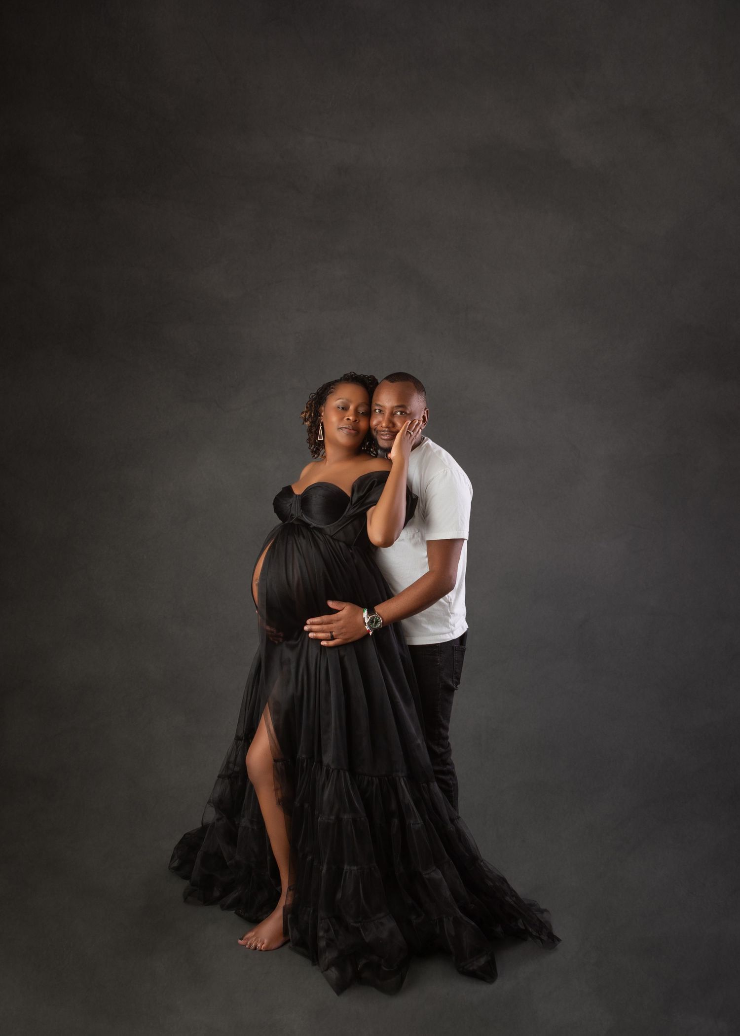 Couple maternity portrait with pregnant woman in black tulle gown embraced by partner on gray backdrop.