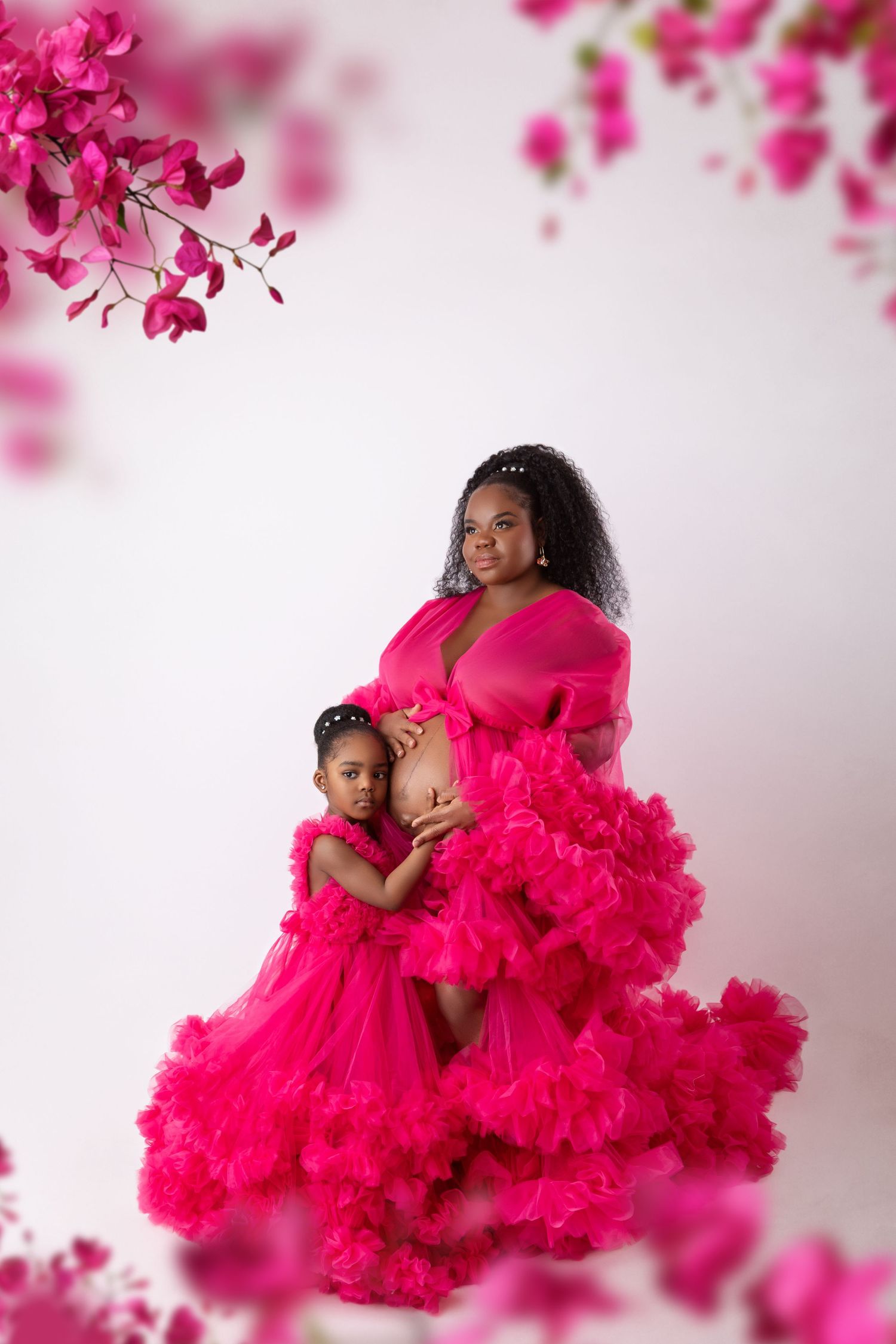 Pregnant woman in hot pink ruffled gown poses with young daughter in matching dress, white background.