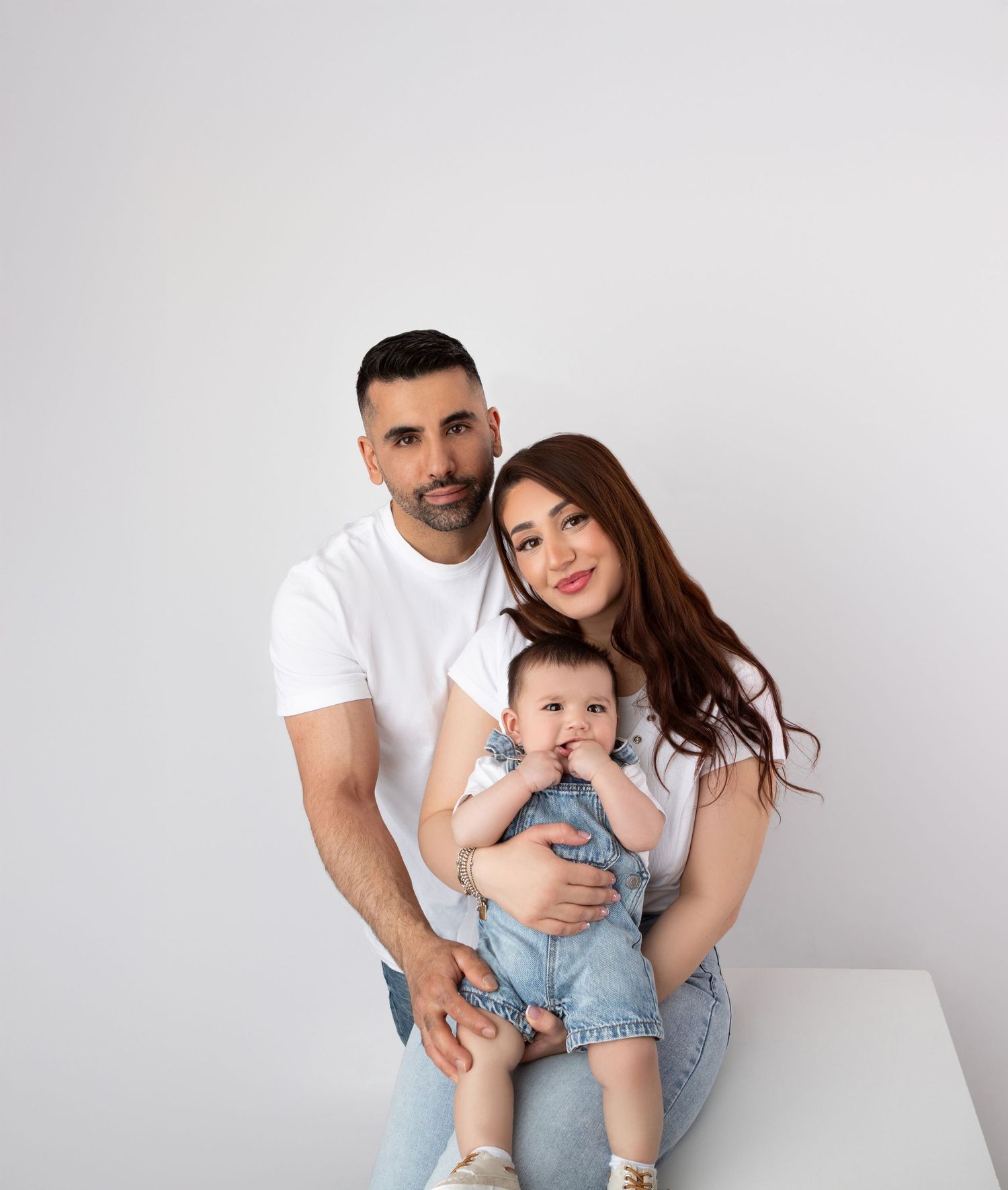 Smiling couple in white tops sits together holding their baby in a bright white studio.