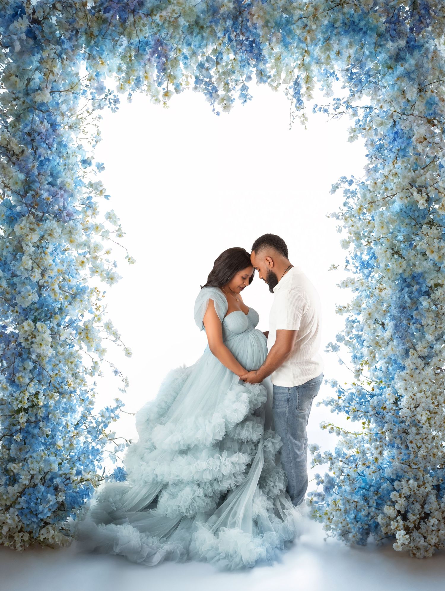 A couple tenderly holds a baby bump under a blue and white floral arch in a maternity portrait.