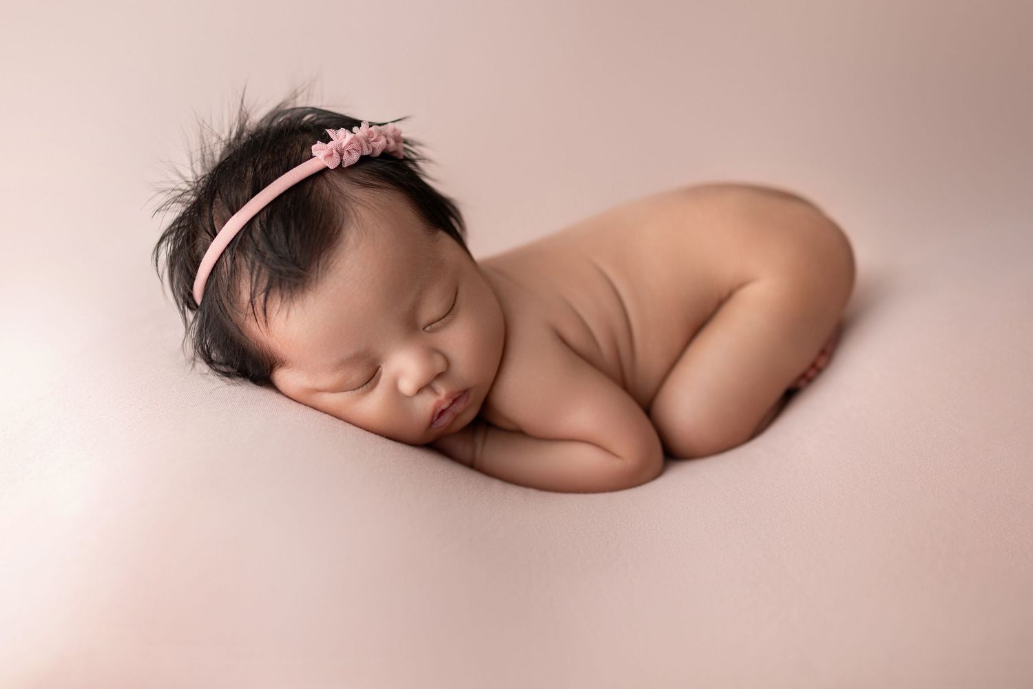 Peaceful newborn baby girl sleeping curled up on pink backdrop wearing a delicate floral headband.
