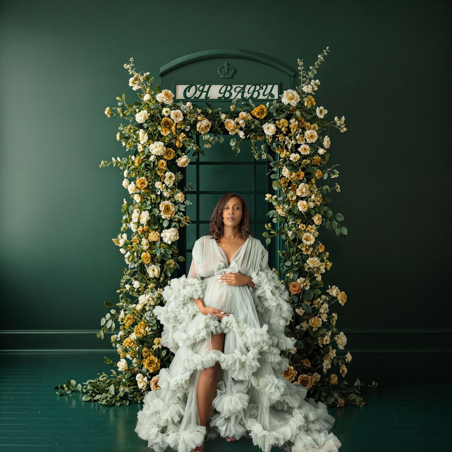 Pregnant woman in white ruffled gown poses under floral arch with yellow roses on green backdrop.