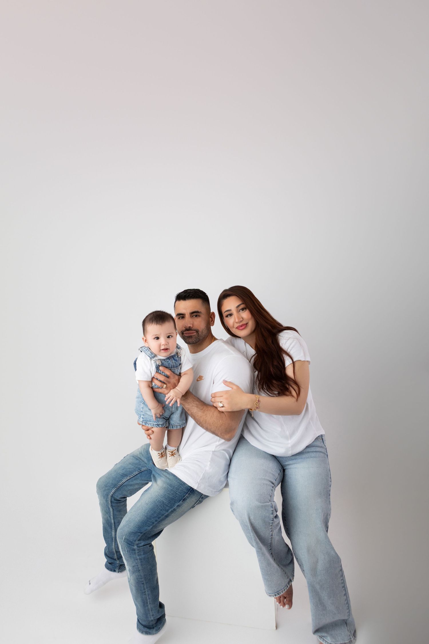 Happy young family with baby in white tees and jeans posing together against a white background.