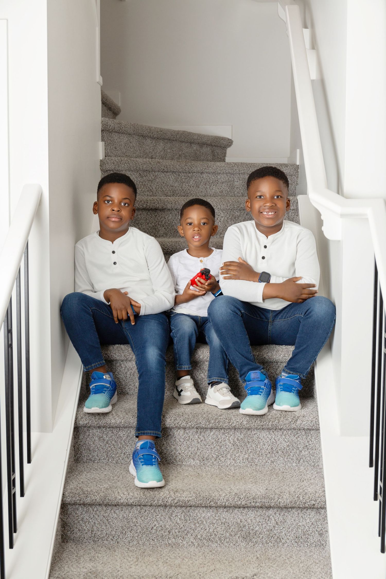 Three young boys in white shirts and jeans sitting together on carpeted staircase smiling at camera.
