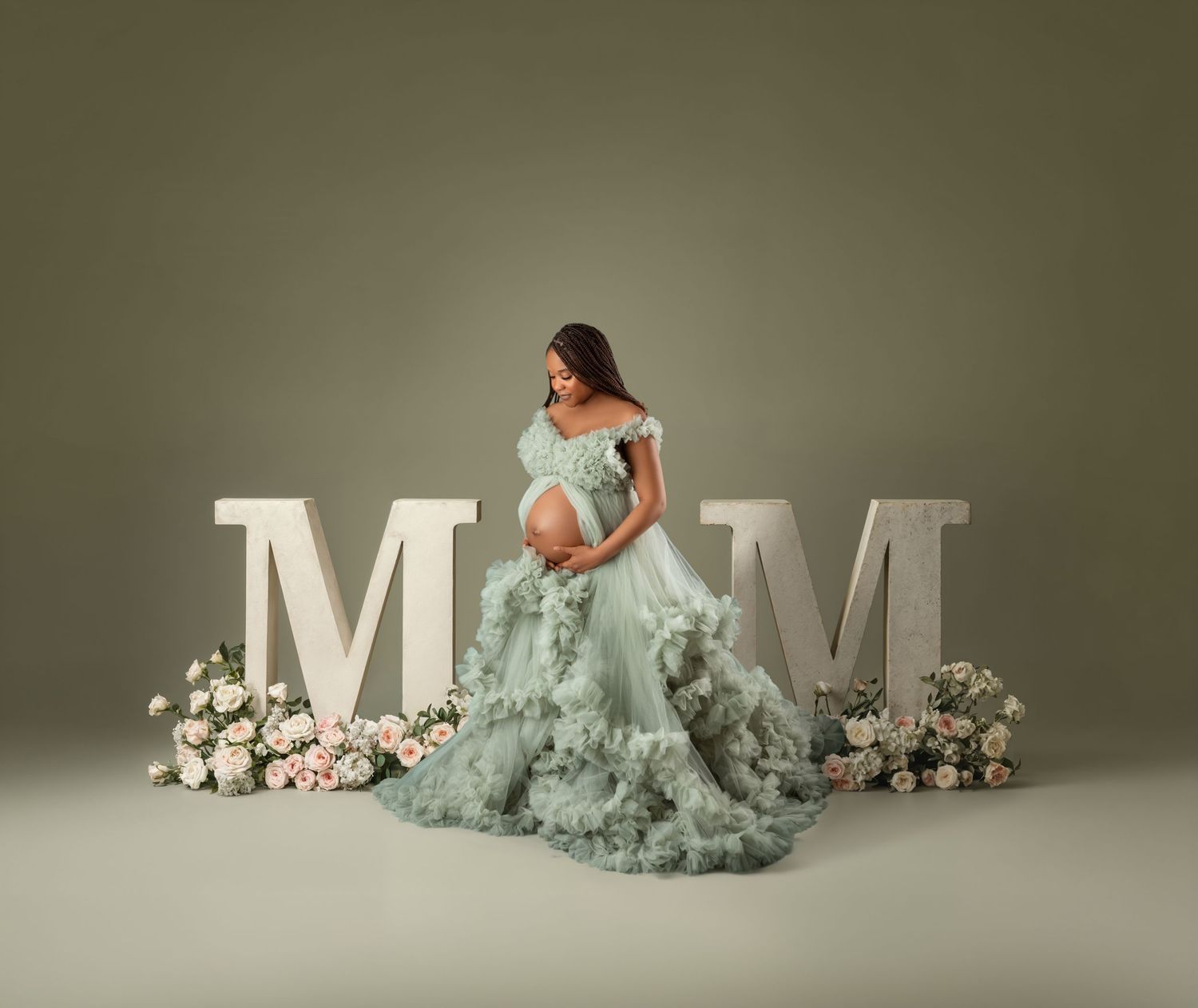 Pregnant woman in sage green ruffled gown poses between large MOM letters with floral arrangements in a studio maternity portrait.