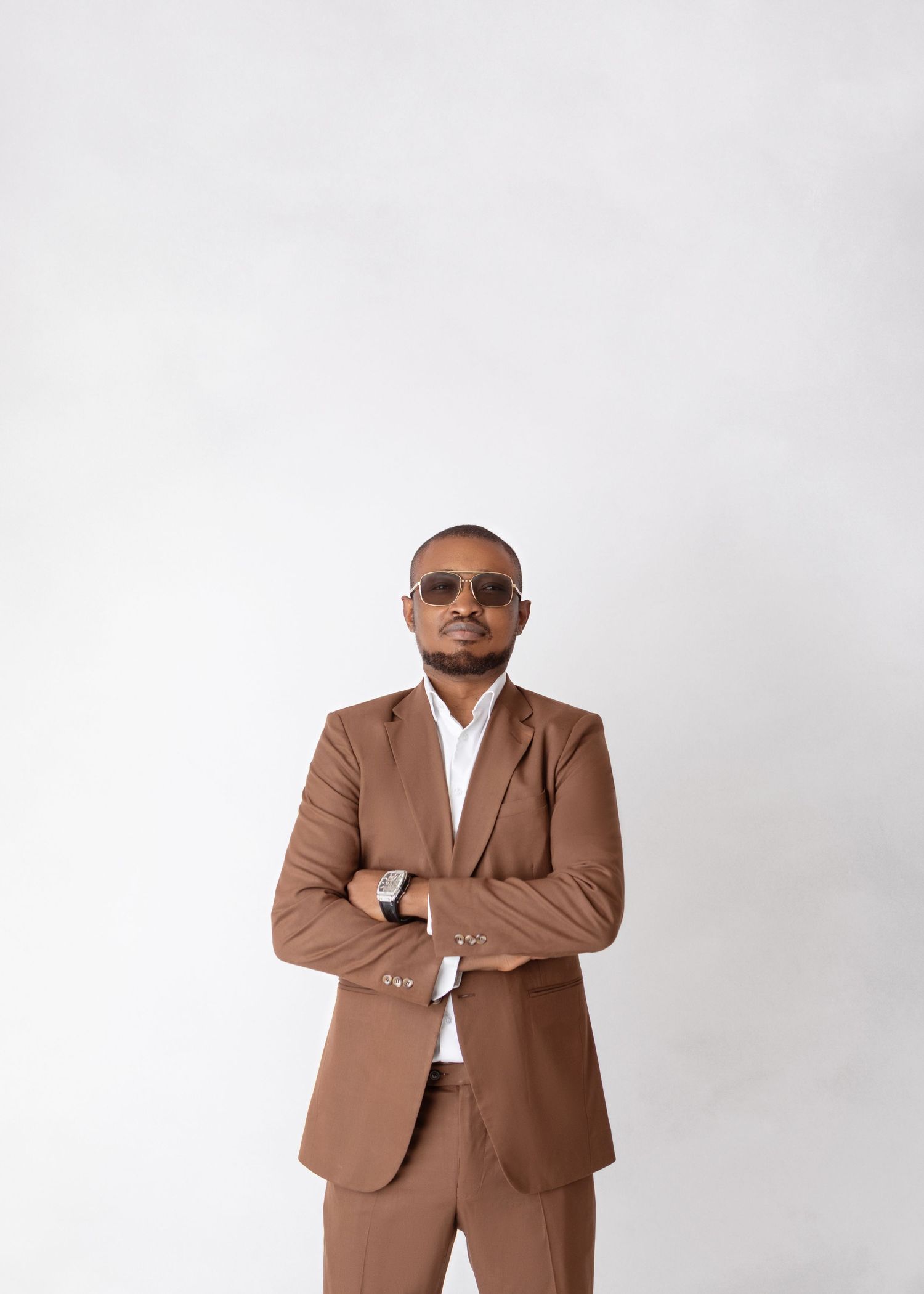 A man in a brown suit with sunglasses stands with arms crossed against a white background.