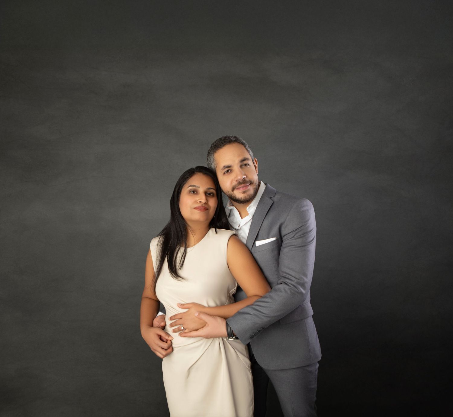 A couple poses intimately against a dark background, woman in cream dress, man in gray suit embracing her.