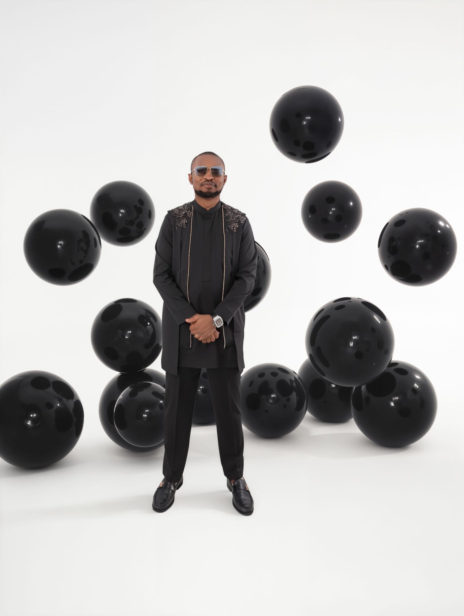 Man in black outfit stands confidently among floating black balloons against white background.