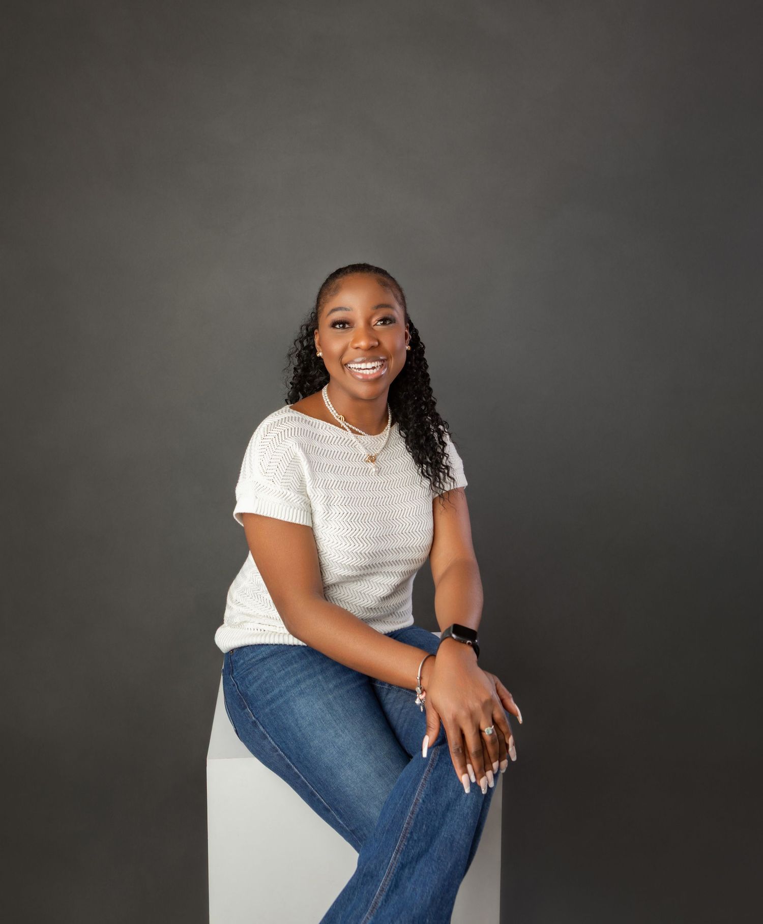 Smiling young woman in white t-shirt and jeans sitting on white cube against dark gray studio background.