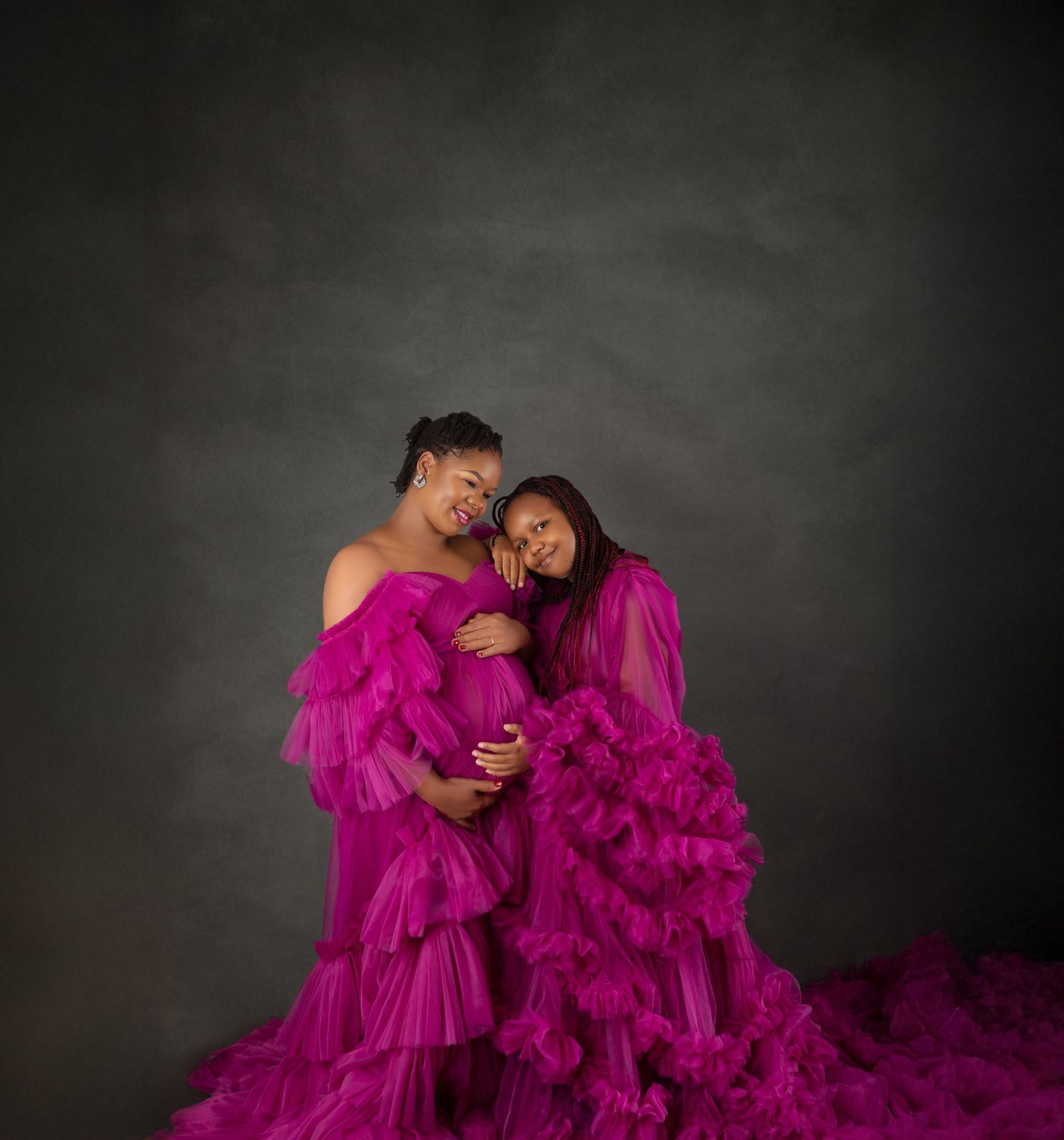 Two women in matching magenta ruffle gowns pose together in a maternity photo shoot against a gray backdrop.