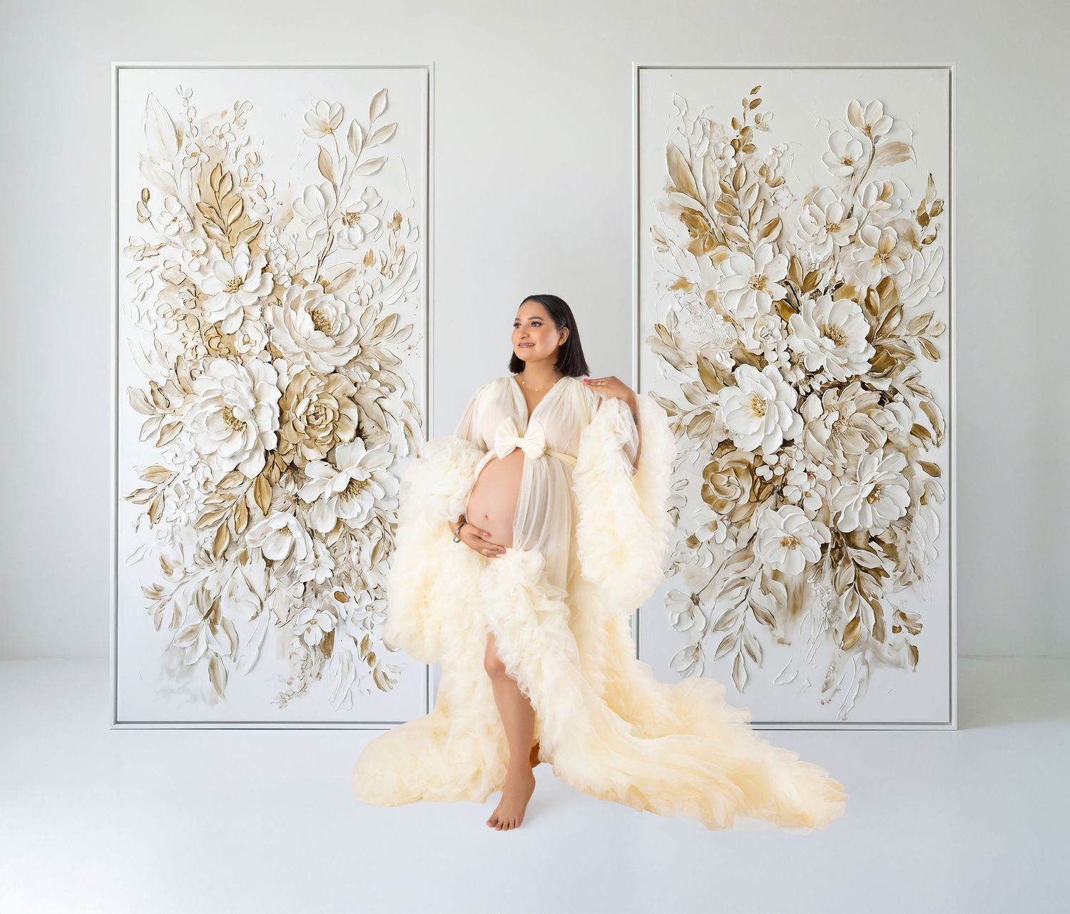 Pregnant woman in elegant cream feather robe gown poses before ornate white and gold floral wall art panels.