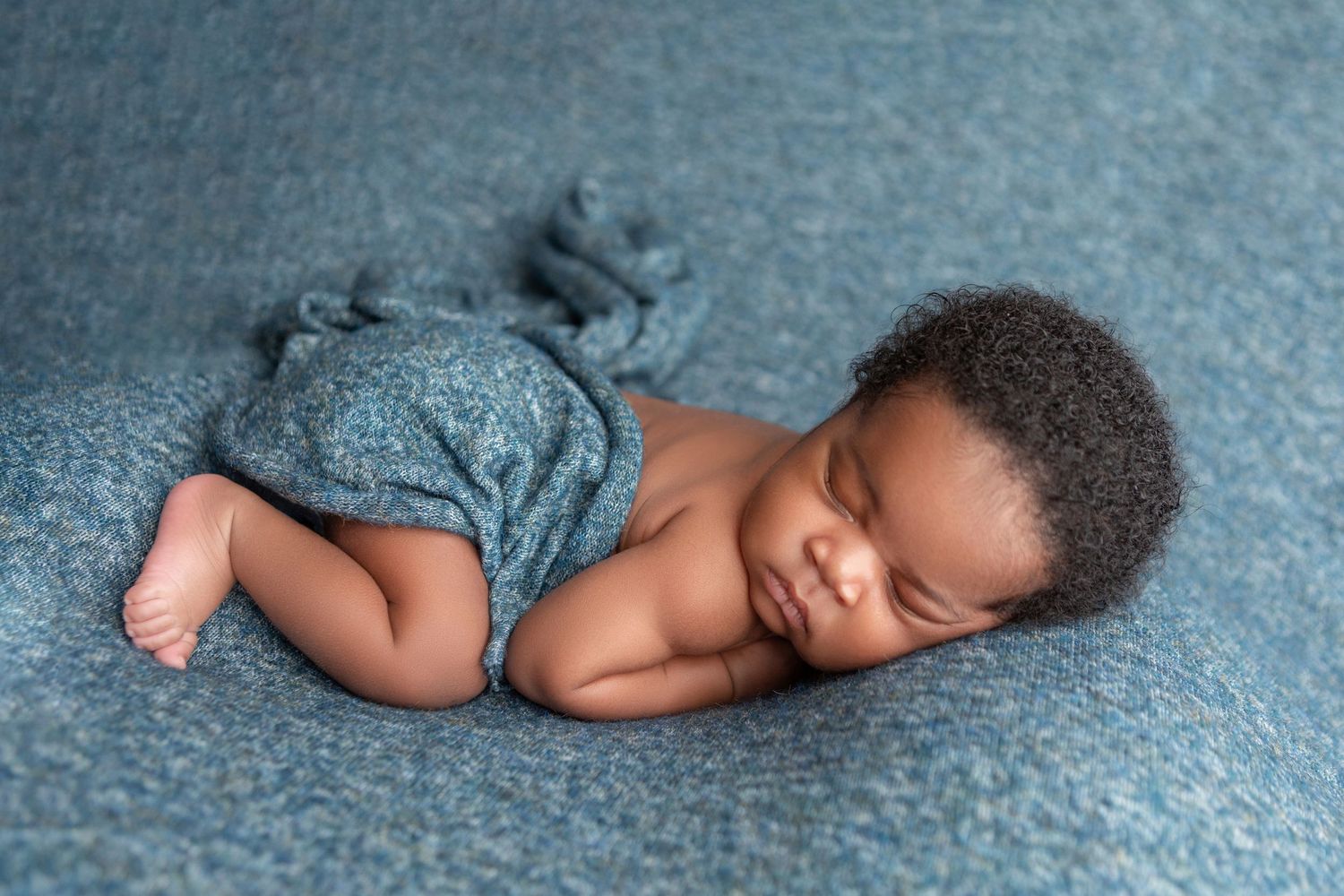 Sleeping newborn baby wrapped in a blue knit blanket on a teal background.