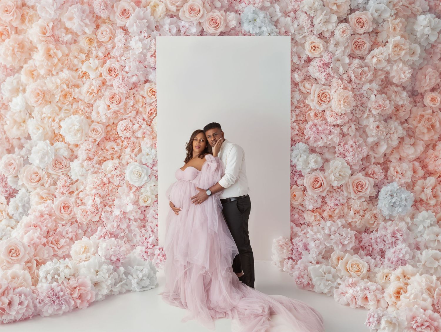 Pregnant couple embraces in pink tulle gown before a lush floral wall in a romantic maternity portrait.