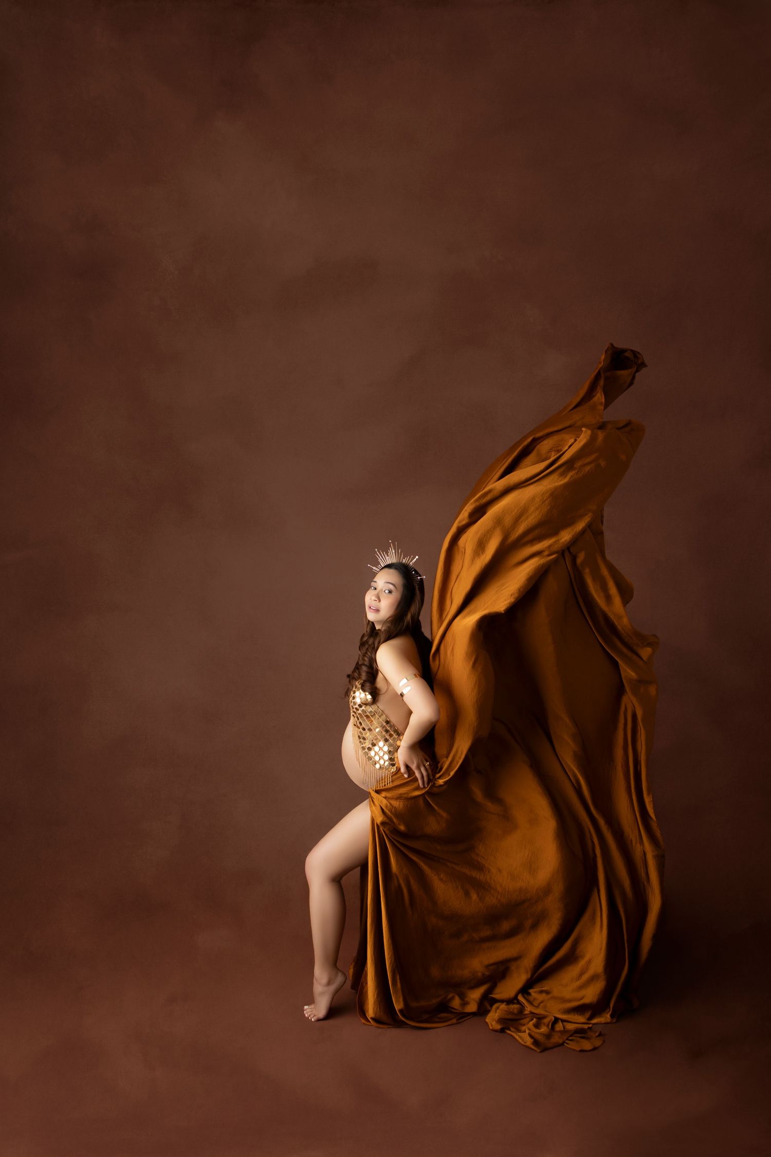 Pregnant woman in golden crown and amber silk fabric billowing dramatically behind her in studio maternity shoot.
