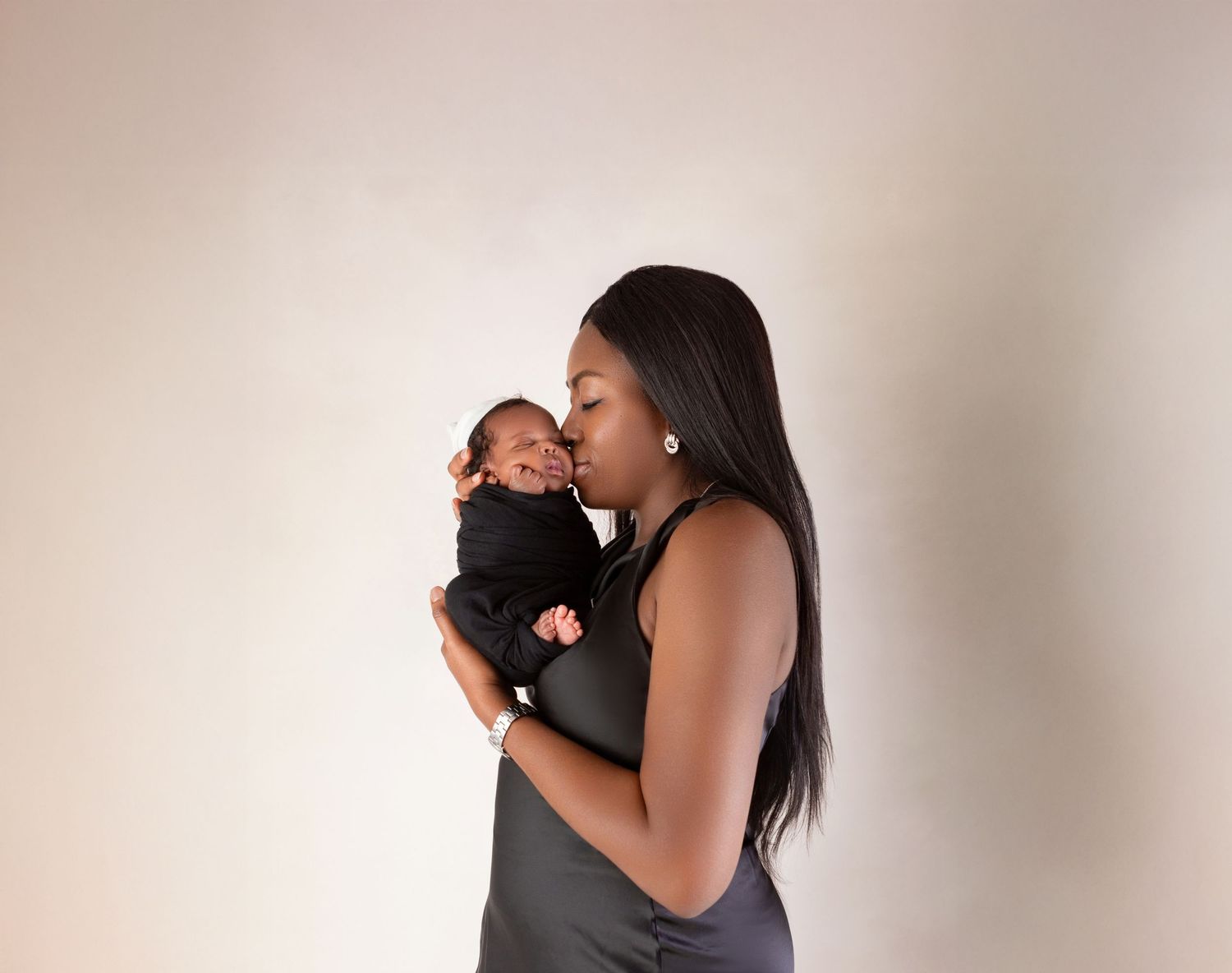 Mother in a black dress gently kisses her swaddled newborn baby against a white wall.