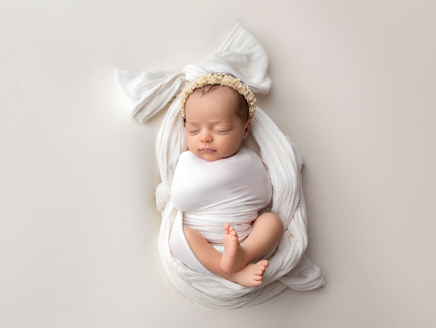 Peaceful newborn baby swaddled in white wrap with floral headband, sleeping on white background.