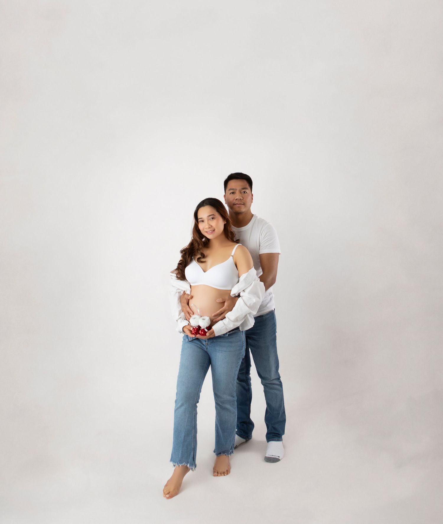 Pregnant couple posing in studio, woman in white bra and jeans, man behind her, both smiling warmly.