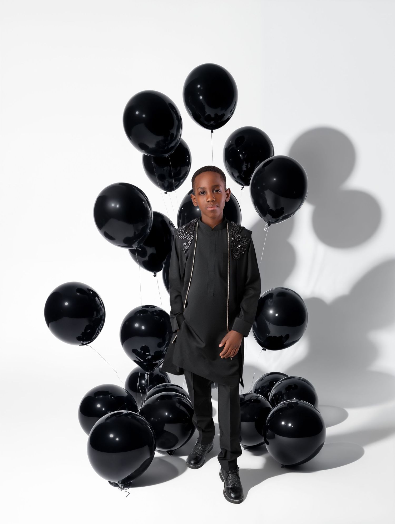 Young person in all-black outfit surrounded by black balloons against white background.
