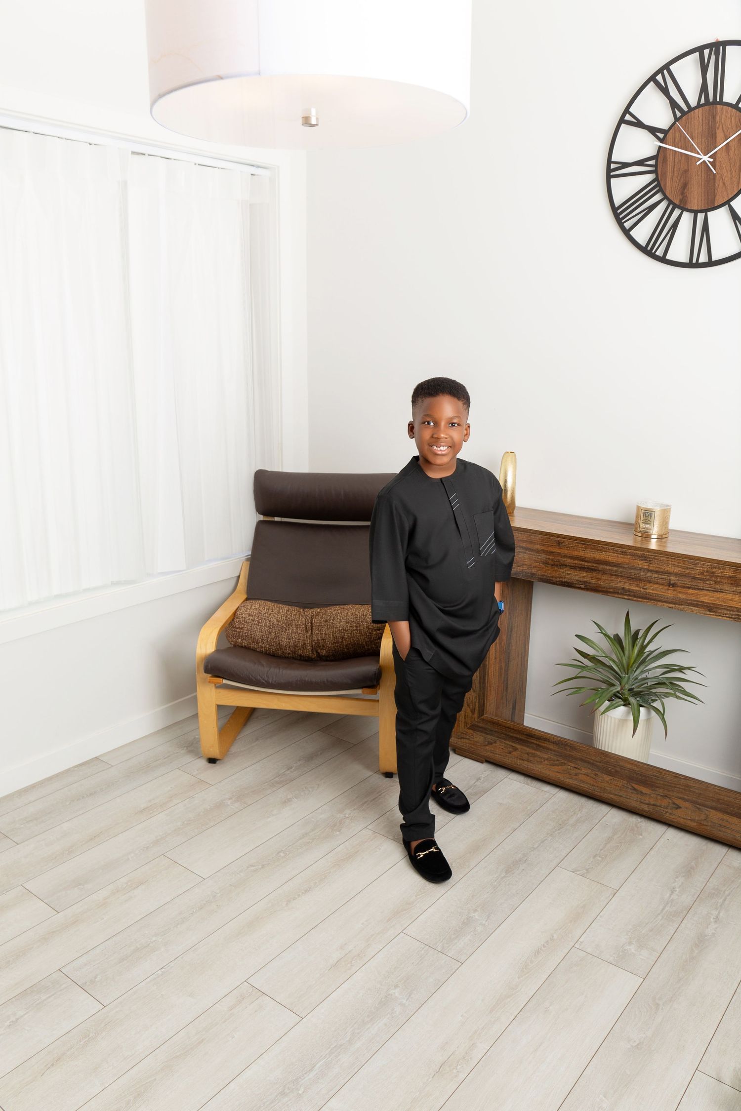 Smiling boy in black traditional attire poses in modern living room with wooden chair, clock, and plant decor.
