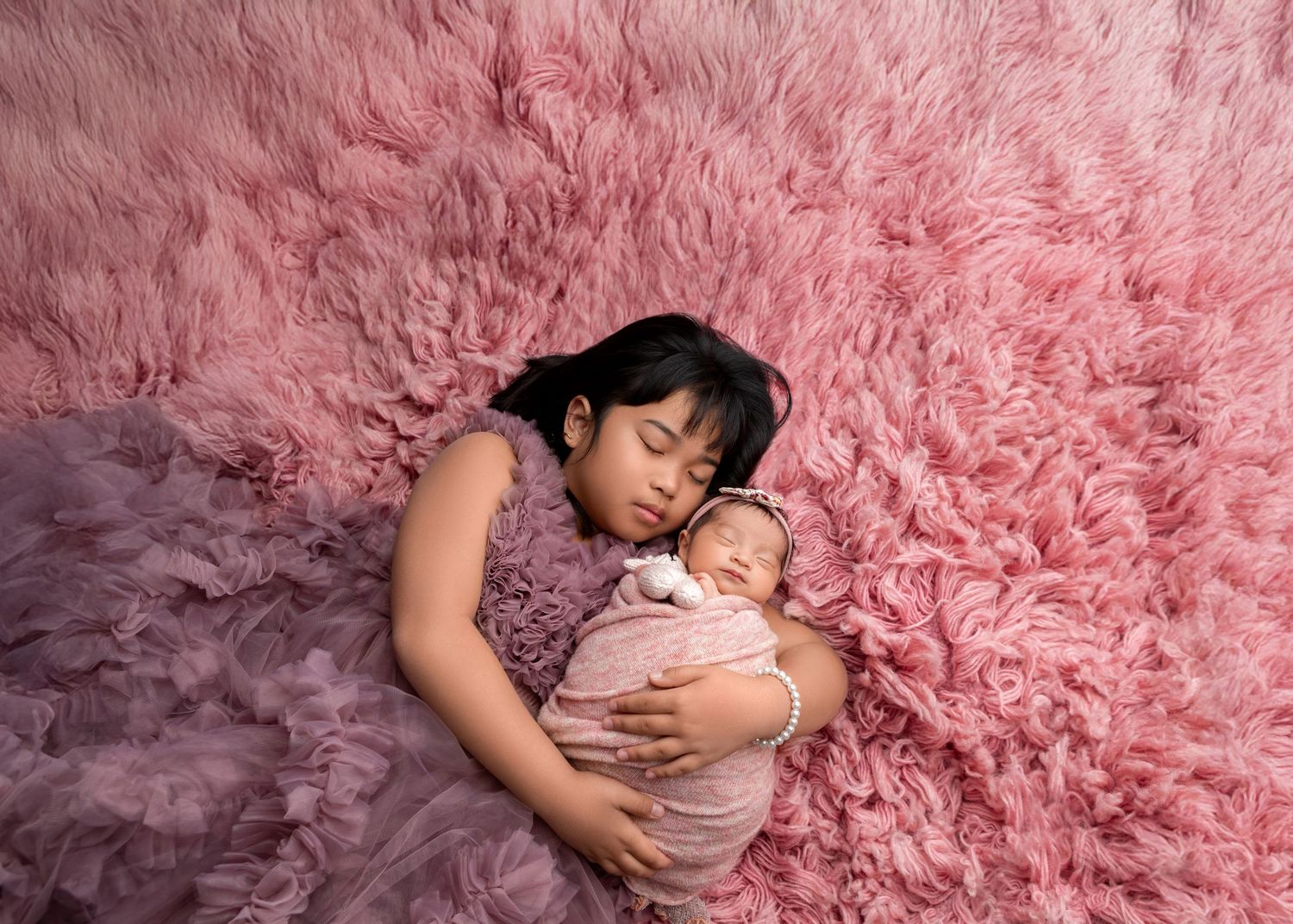 Young girl cuddles sleeping newborn baby on pink fluffy rug in adorable sibling photo.