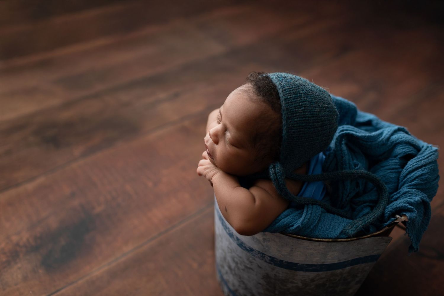 Sleeping newborn baby in teal knit hat nestled in rustic metal bucket with blue wrap on wood floor.