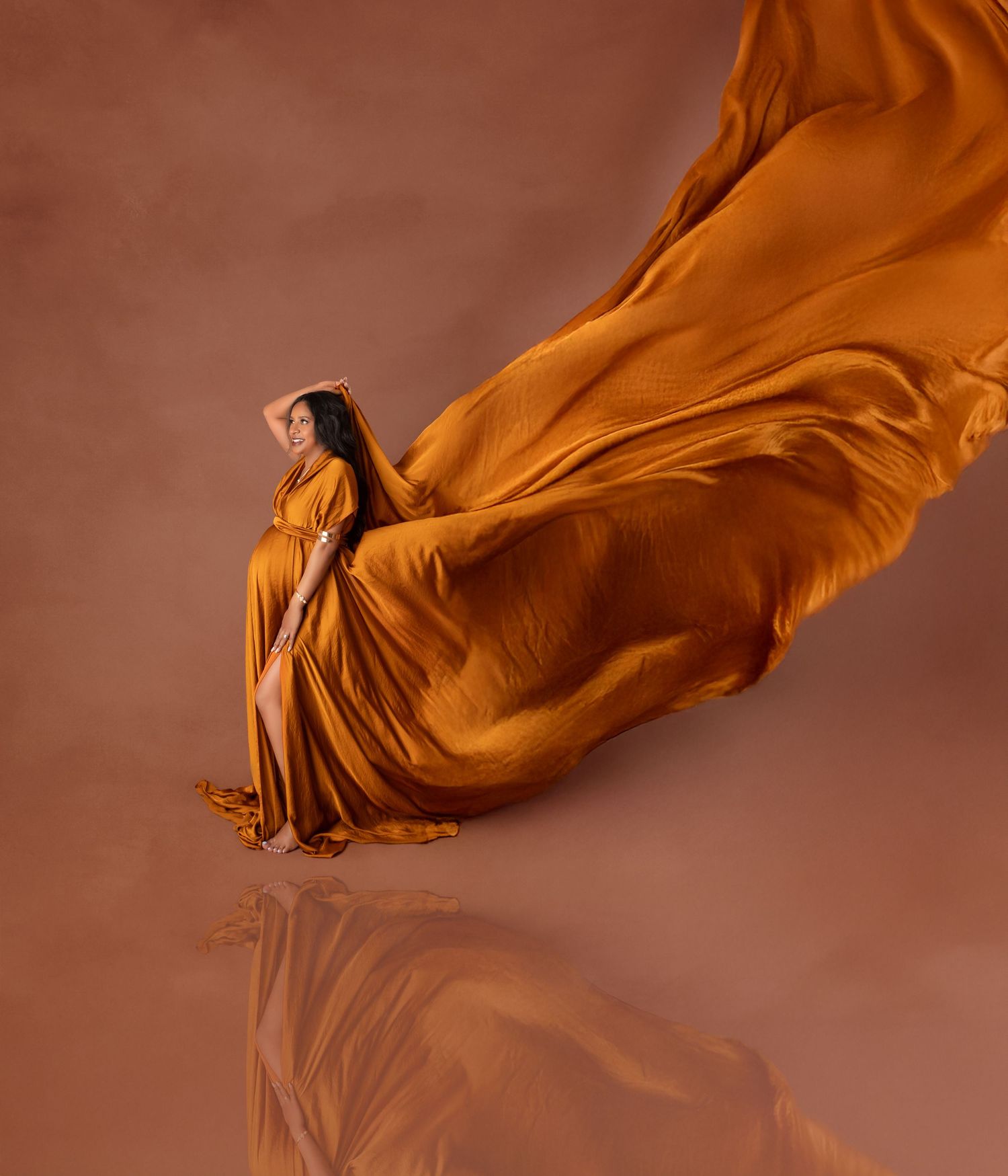 A pregnant woman in a flowing golden orange gown with billowing fabric poses in a reflective studio.
