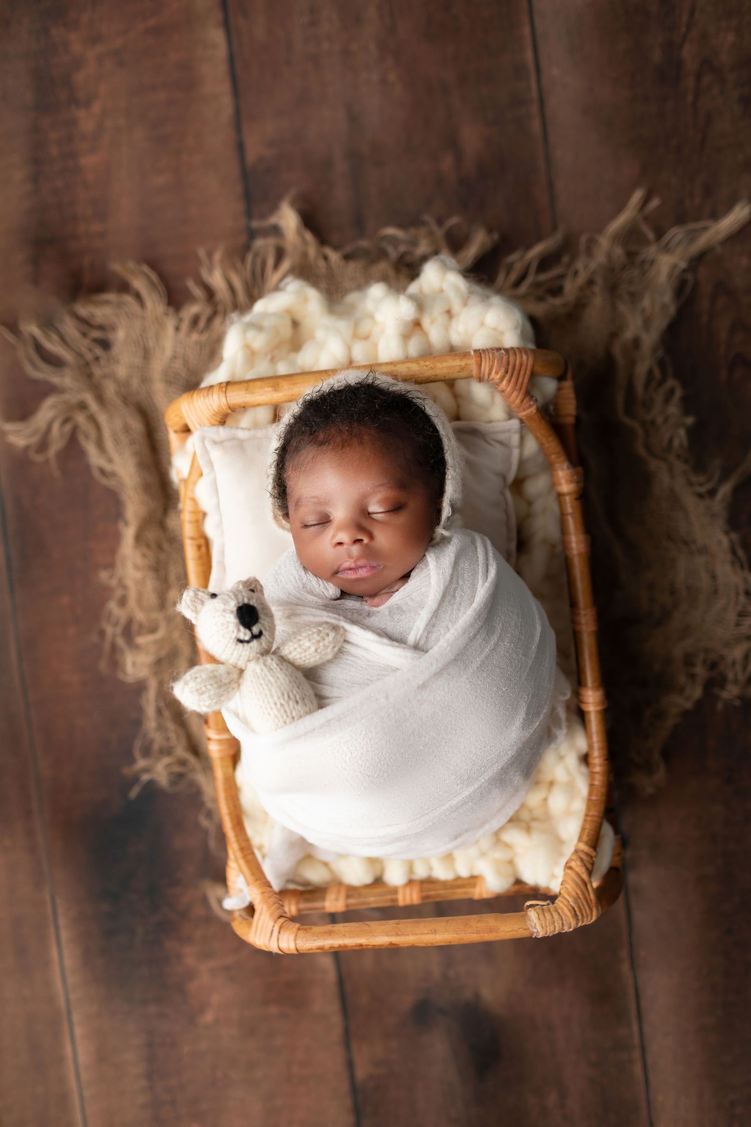 Newborn baby swaddled in white wrap holding a teddy bear, sleeping in a wicker bassinet.