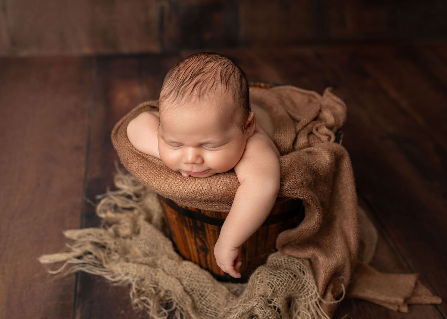 Sleeping newborn baby curled up in a rustic wooden bucket with burlap fabric on a dark wood floor.