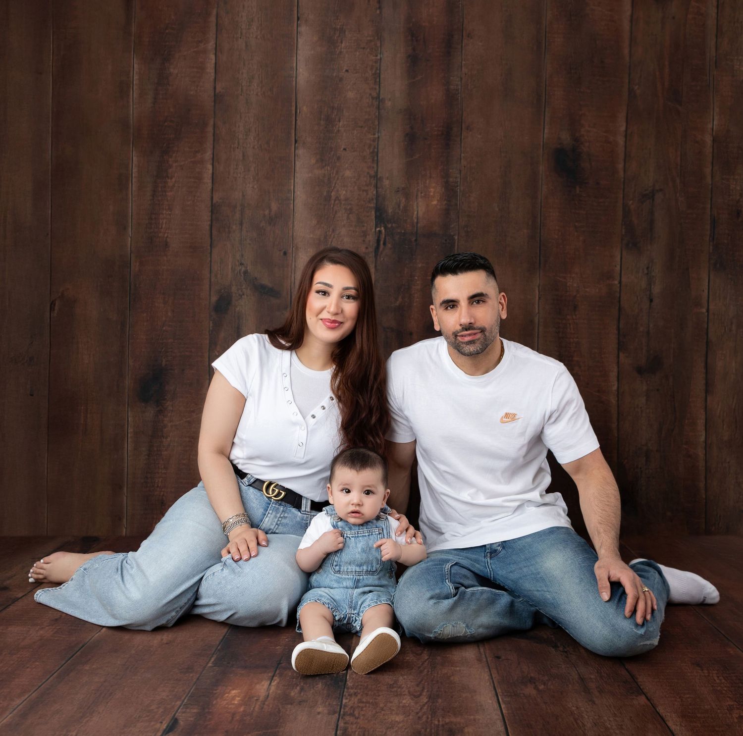 Family portrait of parents and baby in denim overalls against wooden backdrop.