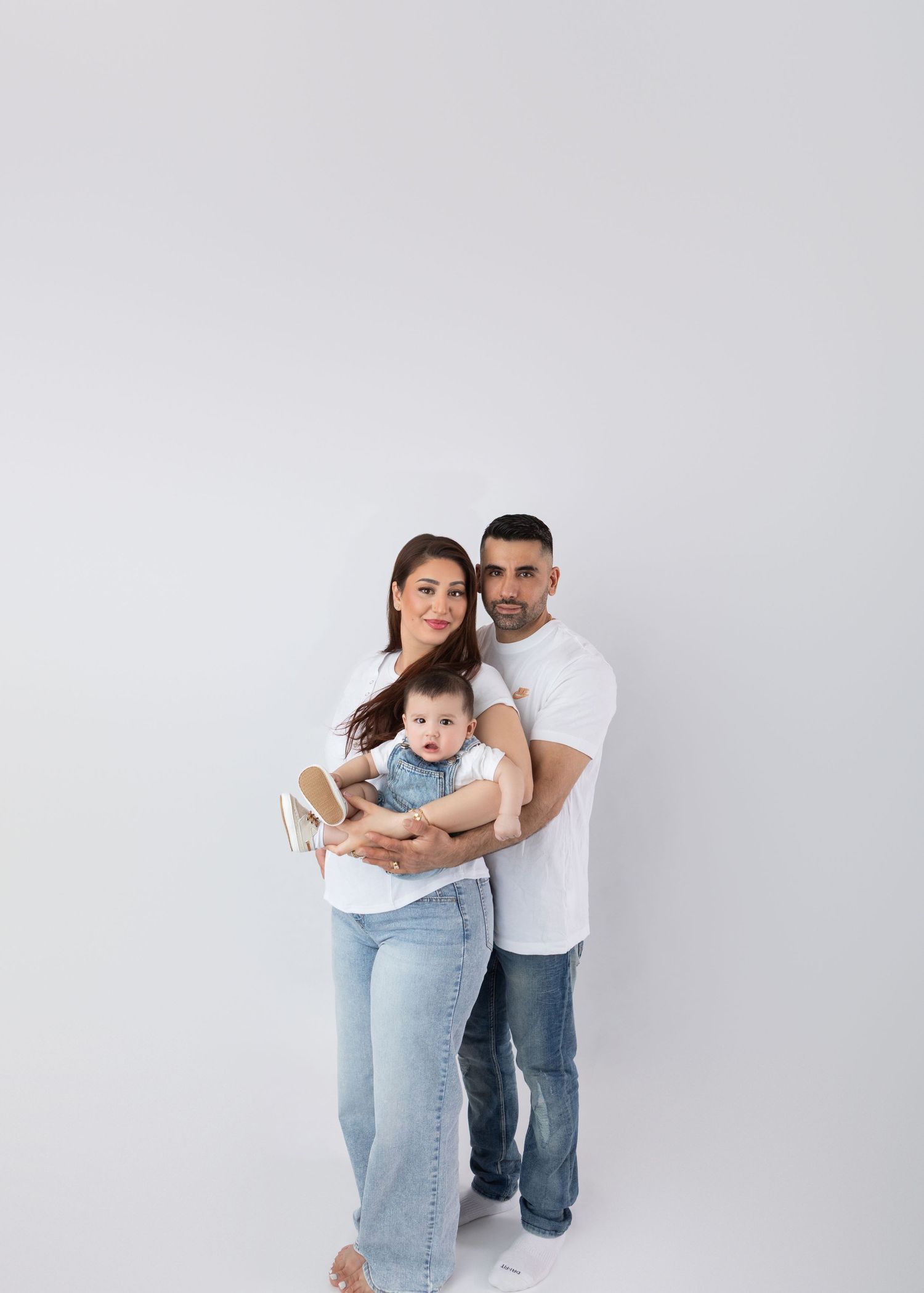 Smiling family of three in white tops and jeans poses in bright white studio.
