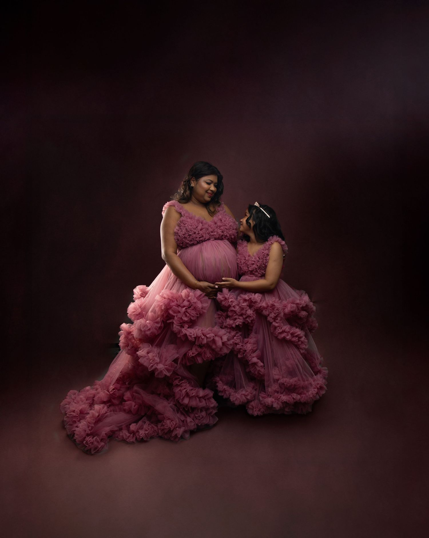 Pregnant mother and daughter wearing matching pink ruffled tulle gowns in a professional maternity photoshoot.