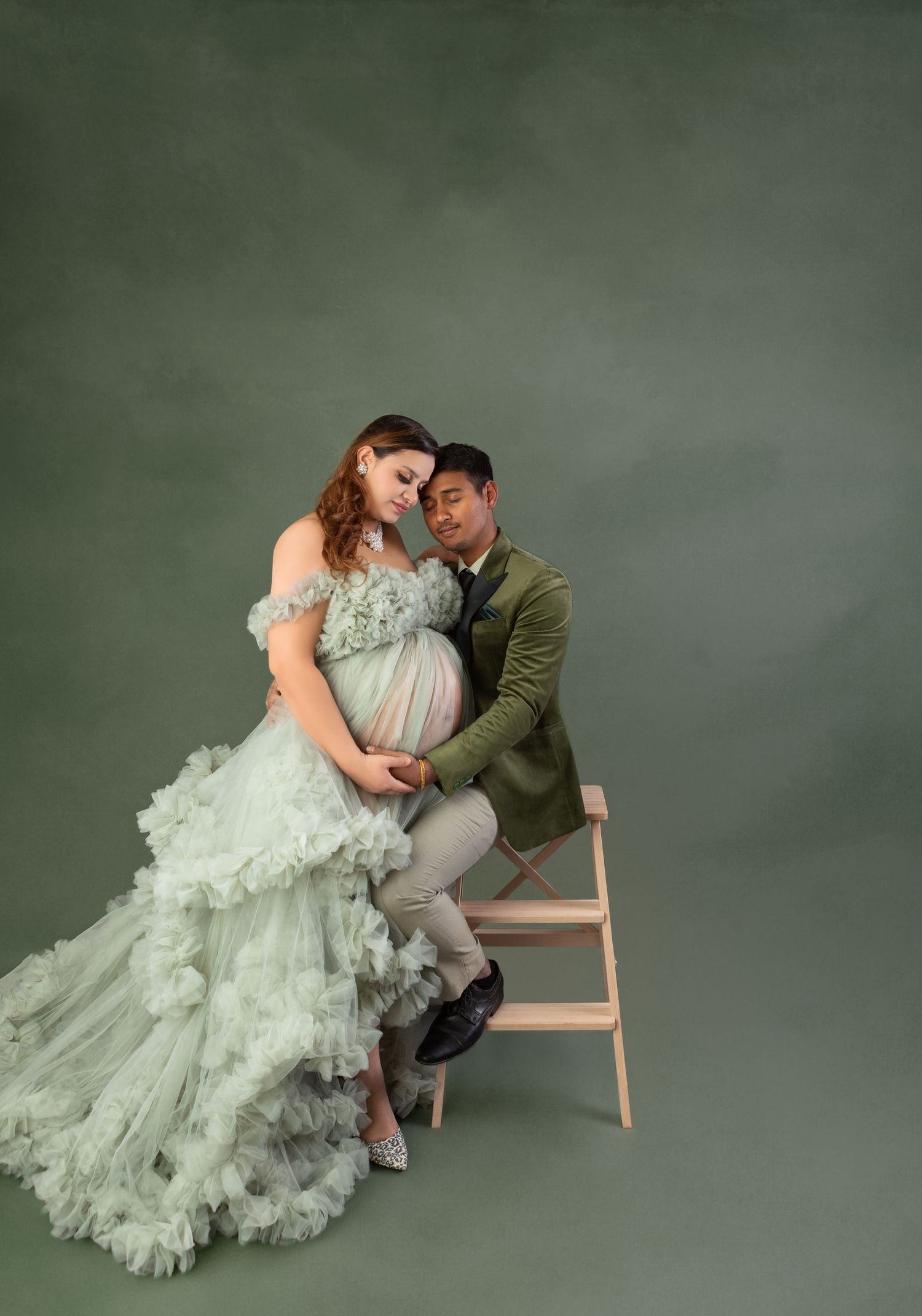 Couple sharing tender maternity photo, woman in green tulle gown, man in olive velvet blazer.
