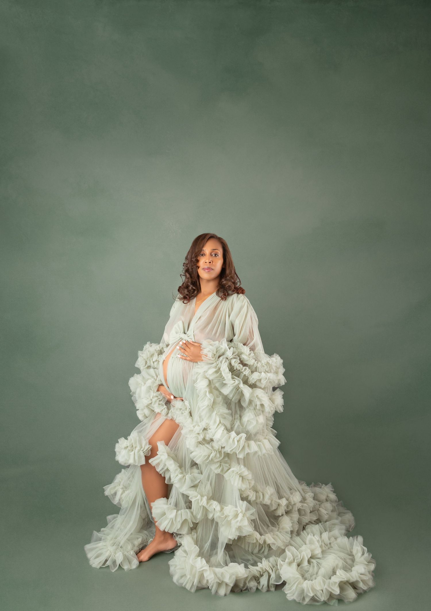 Pregnant woman in white ruffled tulle maternity robe cradles bump while seated against sage green backdrop.