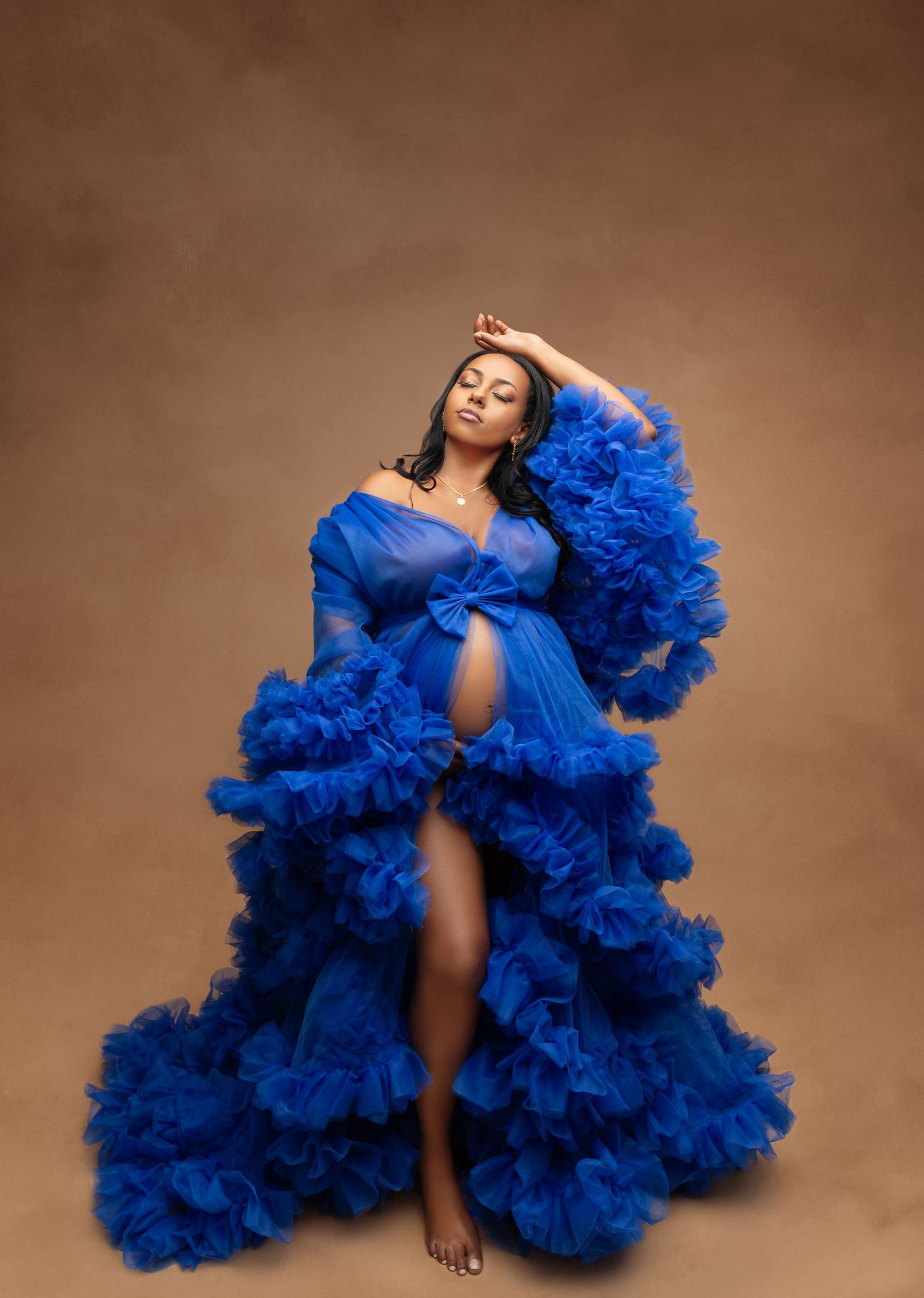 Pregnant woman posing in a stunning royal blue ruffled tulle maternity gown against a warm brown studio backdrop.