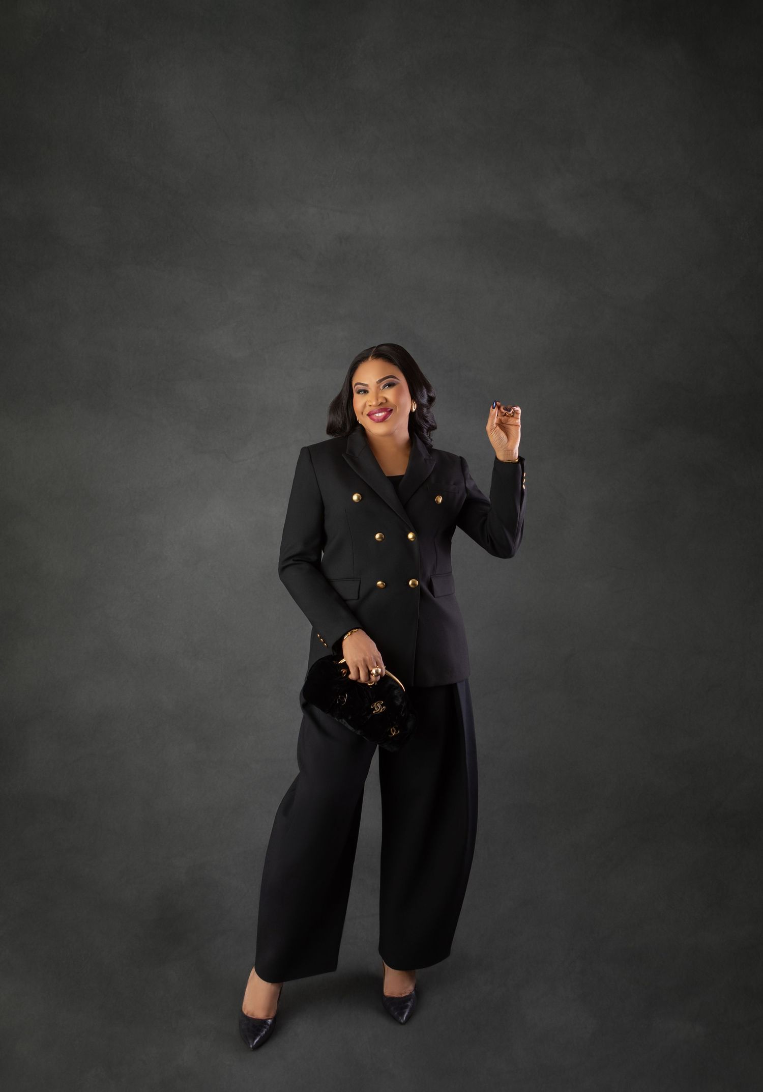 Woman in black double-breasted blazer and wide-leg pants posing confidently with clutch bag on gray background.