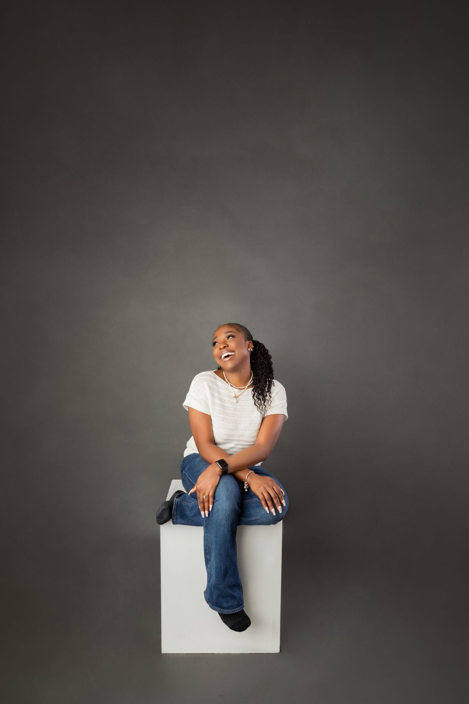 Young Black woman in white tee and jeans sits on white cube, smiling upward against gray studio background.