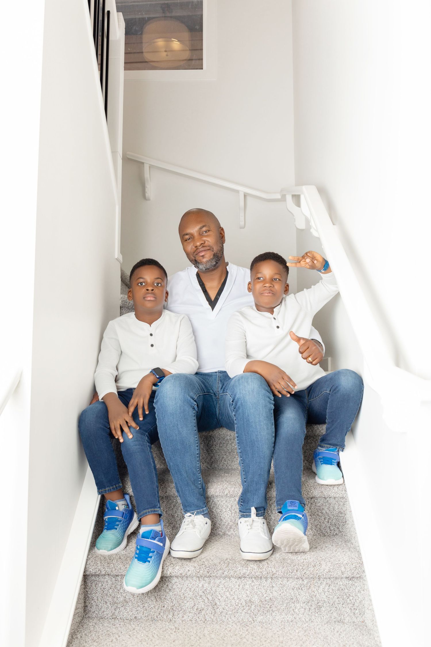A father and his two young sons sit together on carpeted stairs, all wearing white shirts and jeans, smiling at the camera.