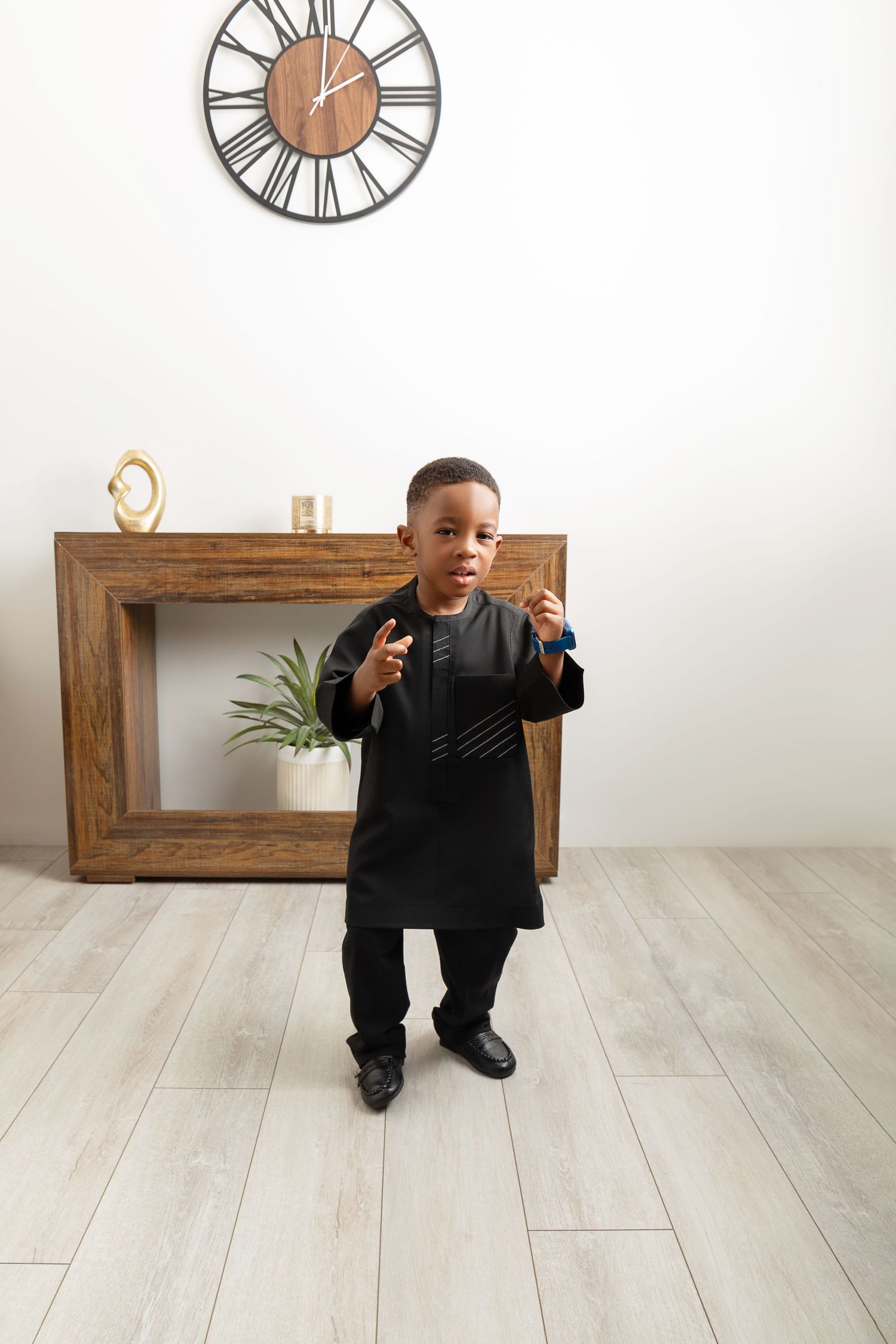 Young boy in black traditional African attire posing confidently in a modern living room with wooden furniture and wall clock.