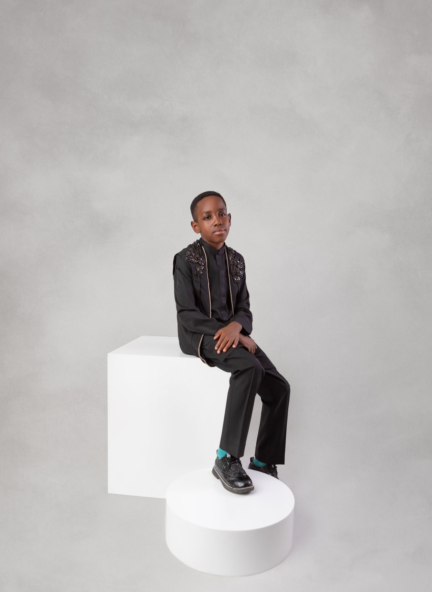 Young boy in black embroidered jacket and trousers seated on white geometric pedestal in photo studio.