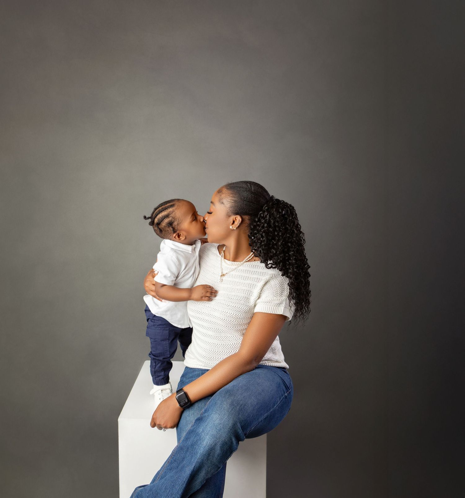 A mother kisses her baby boy on the forehead during a studio portrait session, both wearing white tops.